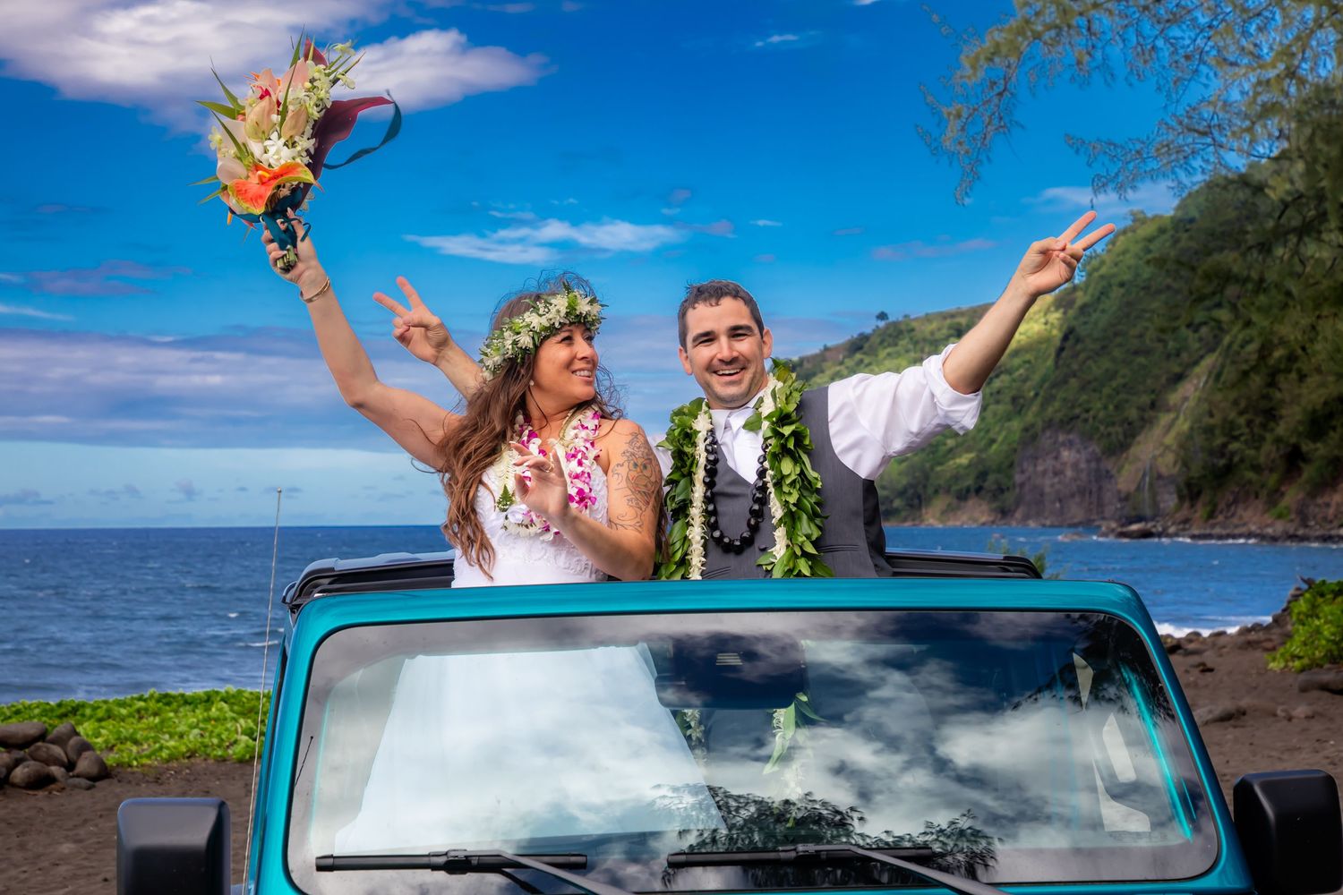 A wedding couple celebrates in a blue Jeep by the Hawaiian coastline with leis and tropical flowers in Waipio Valley on the Big Island.