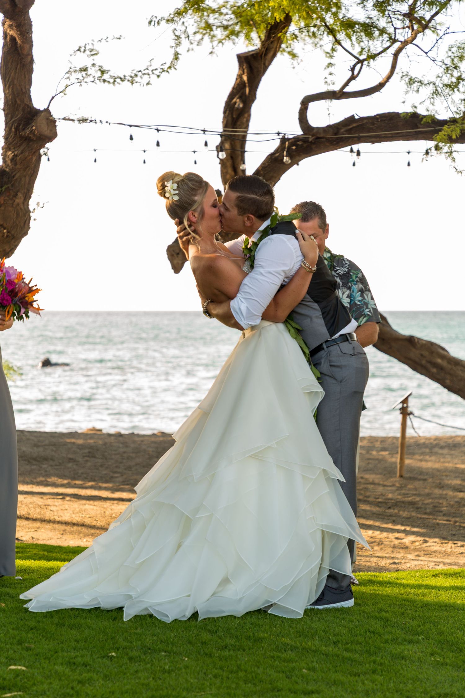 Wedding couple shares romantic dip pose against beach backdrop with lush trees overhead during outdoor ceremony.