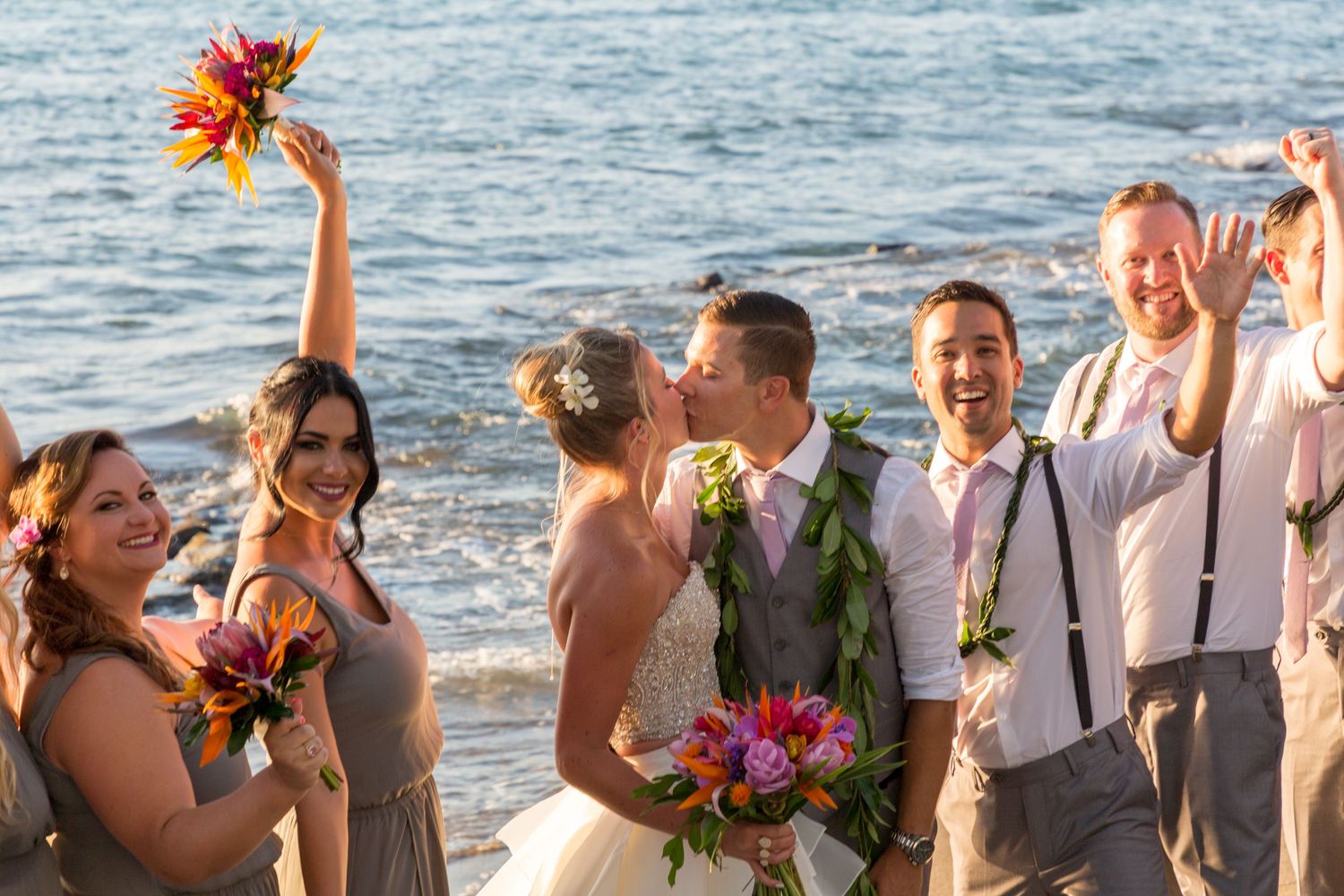 Wedding party celebrates joyfully on a sunny beach with vibrant orange and pink flowers by the ocean.
