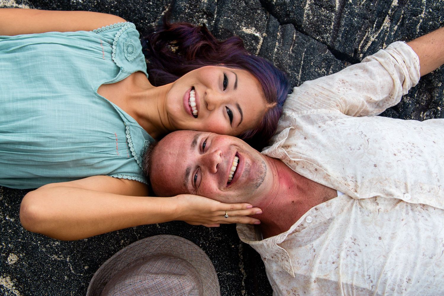 Happy couple lying on lava rock laughing together outdoors during engagement session at Keiki Beach Queen's Bath in Kailua Kona, Hawaii.