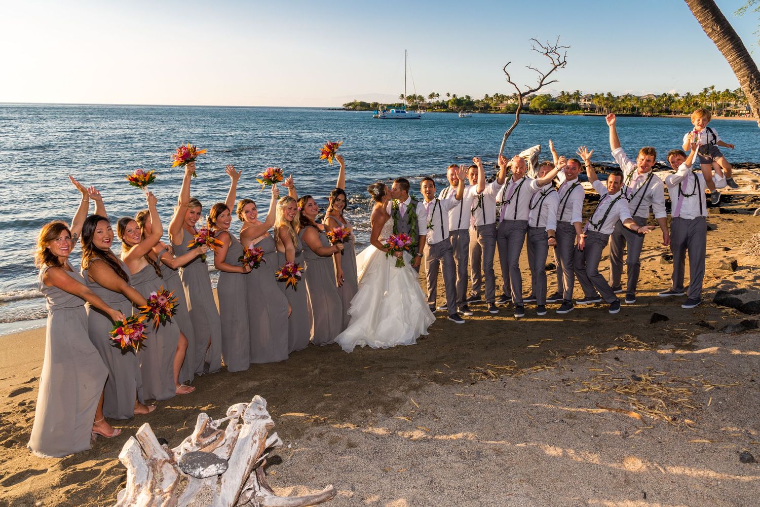 The wedding party celebrates and poses together on a scenic beach at sunset with palm trees and ocean views in the background.