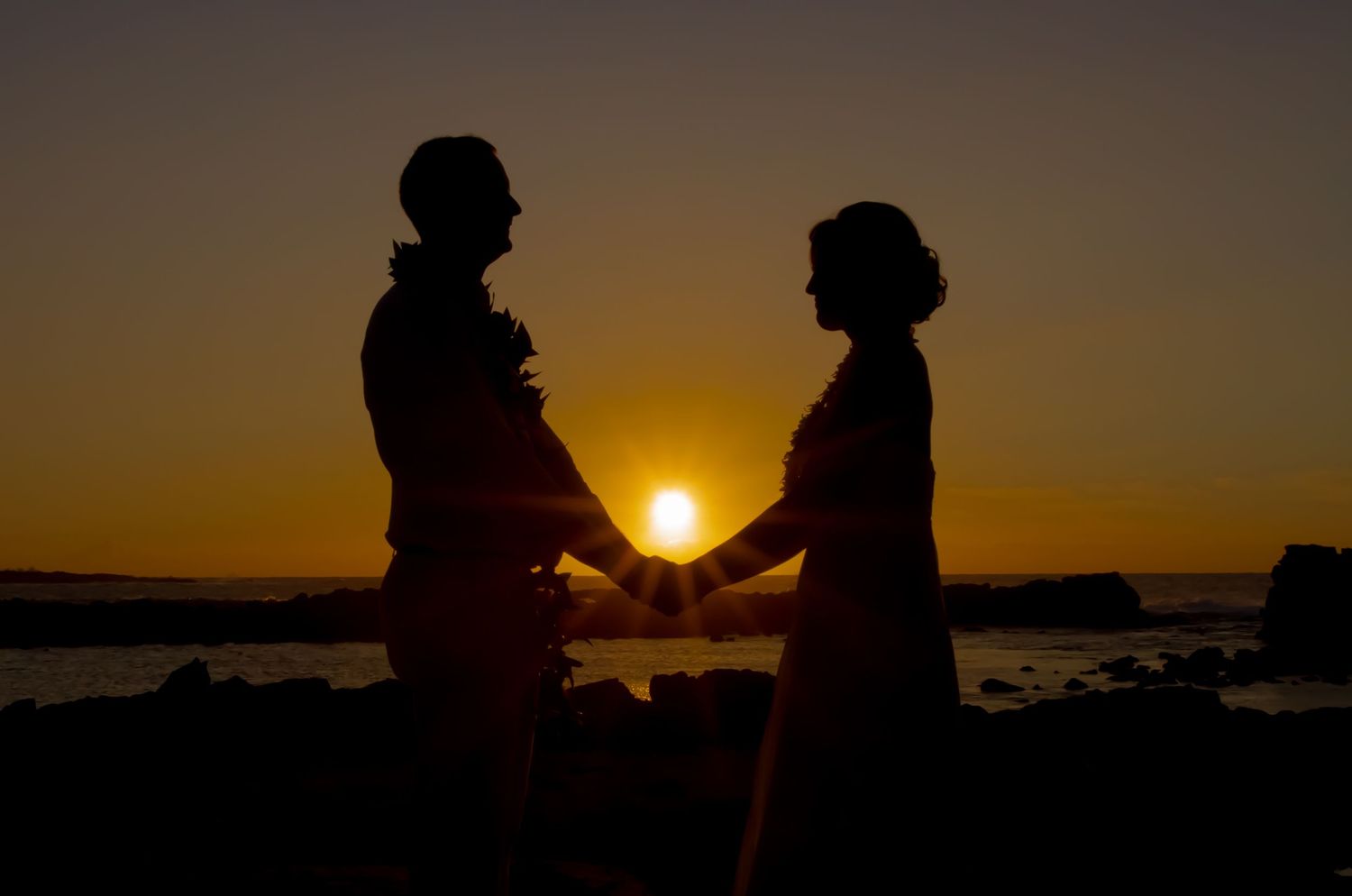 A romantic silhouette of a wedding couple holding hands at sunset with the glowing sun perfectly centered between them.