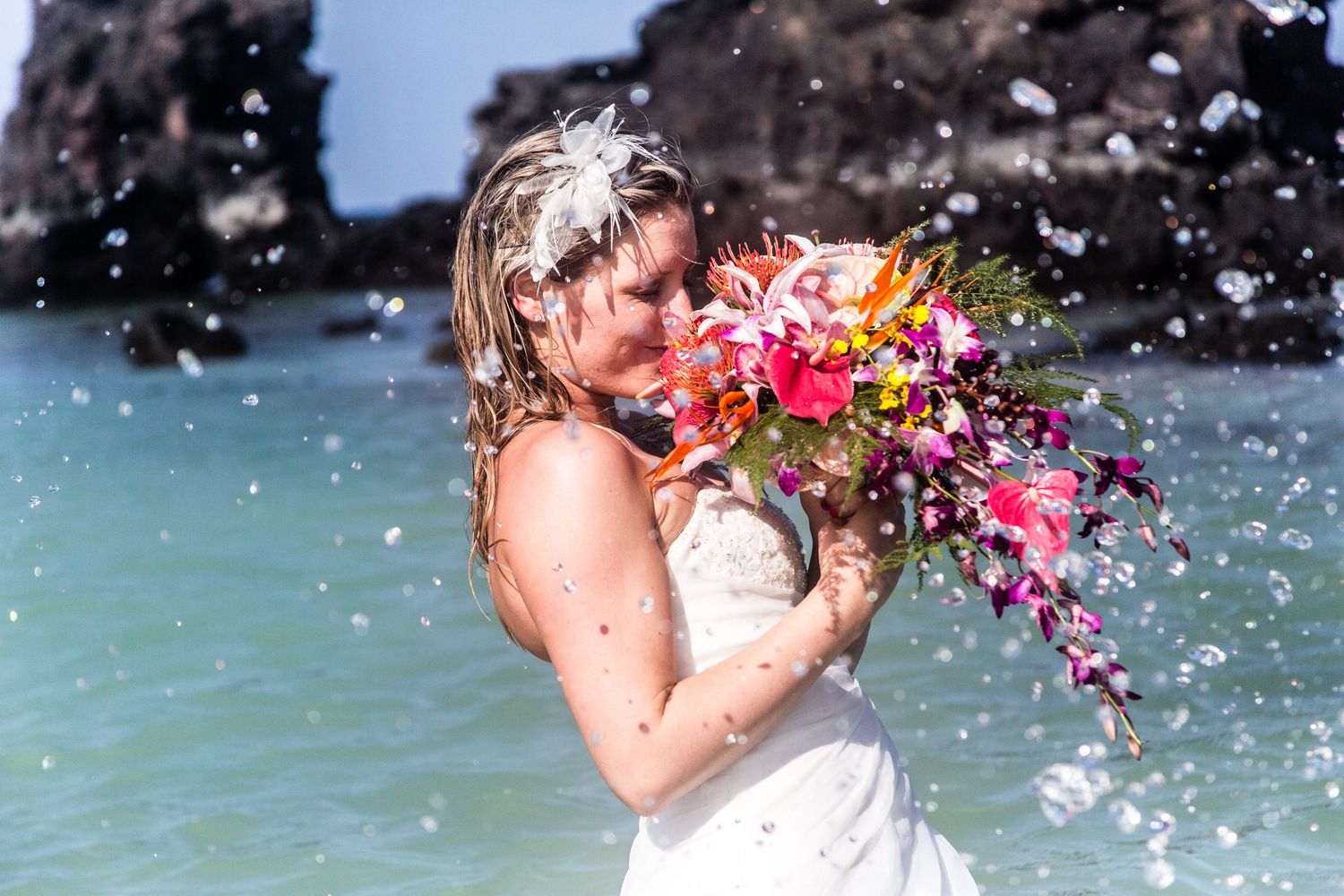 A bride in a white dress holds a vibrant tropical bouquet while ocean waves splash around her on Kikaua beach in Kailua Kona.
