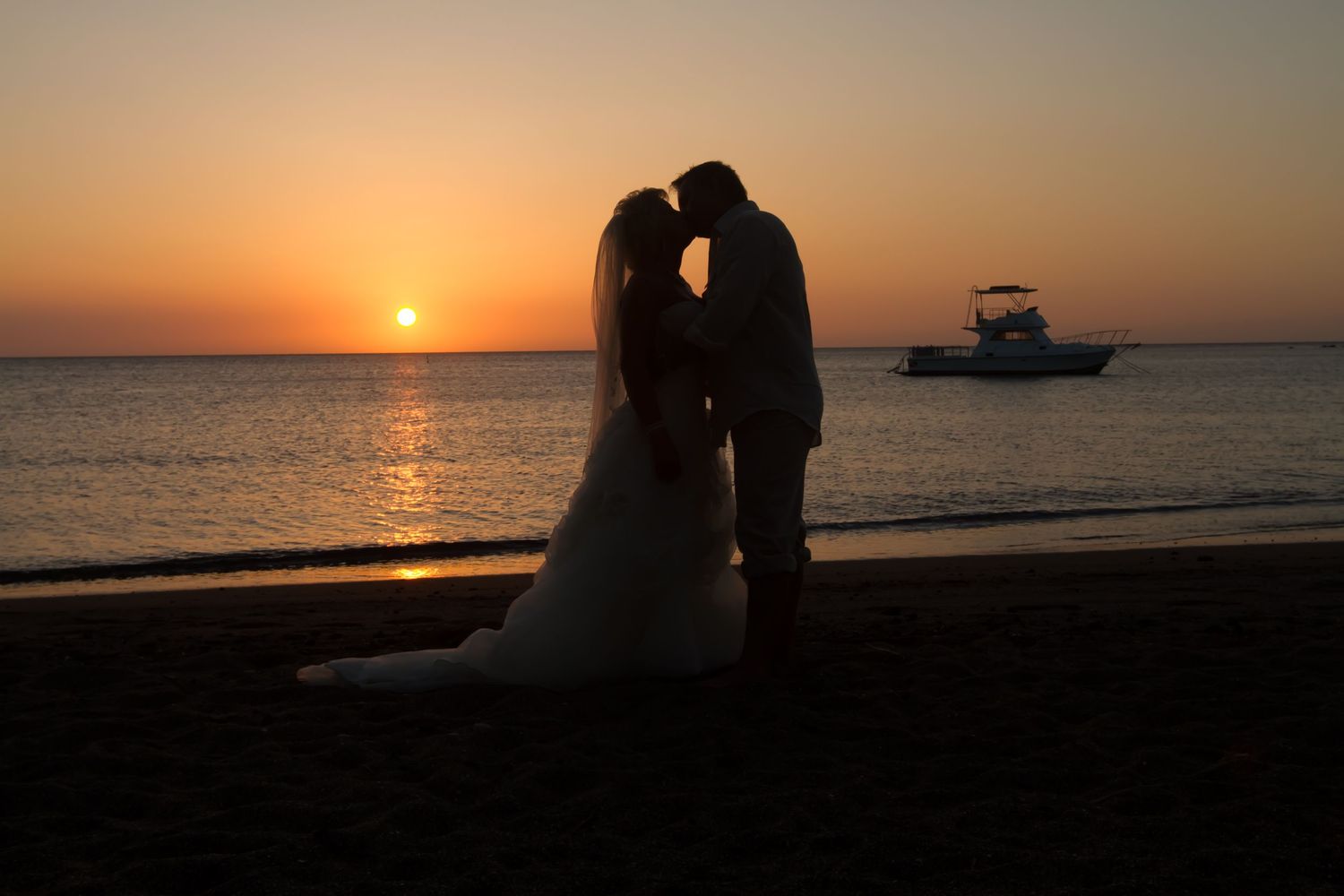 Silhouettes of couple in romantic beach wedding pose at sunset with boat on horizon.