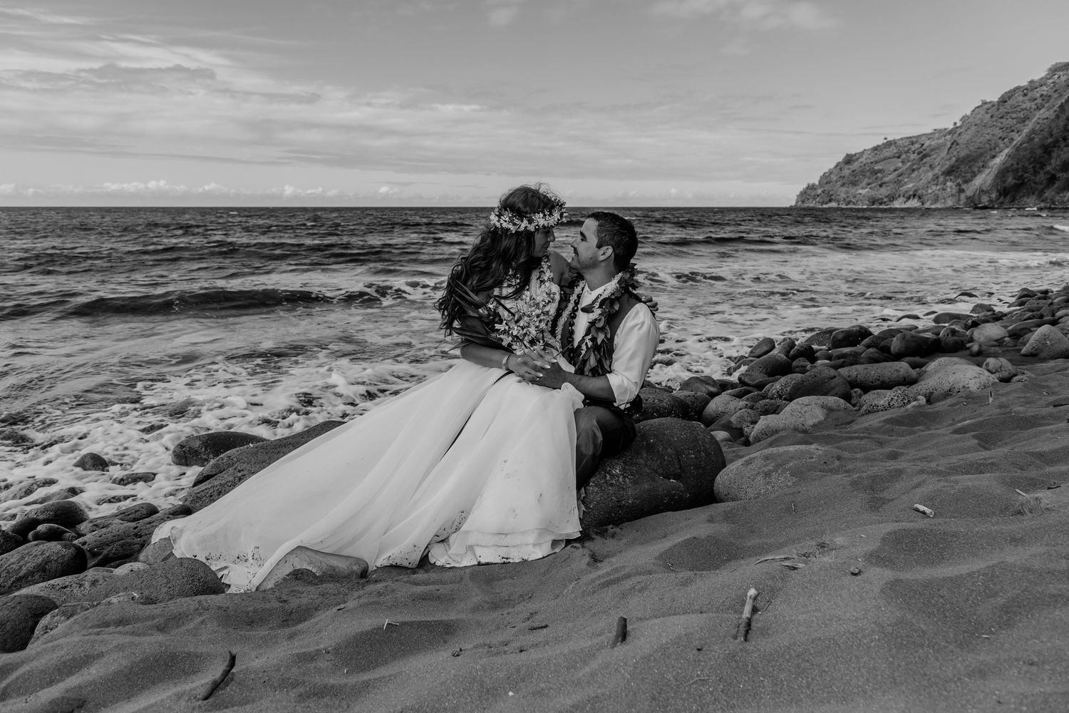 A romantic black and white photograph of a couple sharing an intimate moment on a rocky beach at sunset in Waipio Valley on the Big Island.