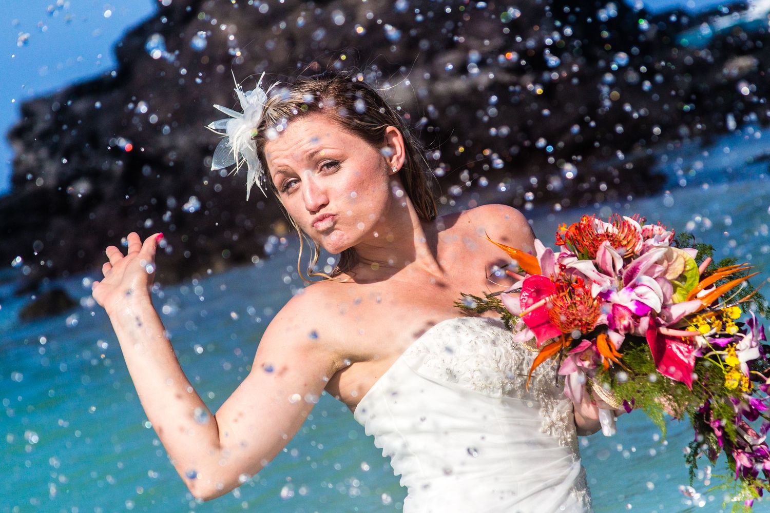 A bride in a white dress holds a tropical bouquet while water droplets sparkle in the bright sunlit air on Kikaua beach in Kailua Kona.