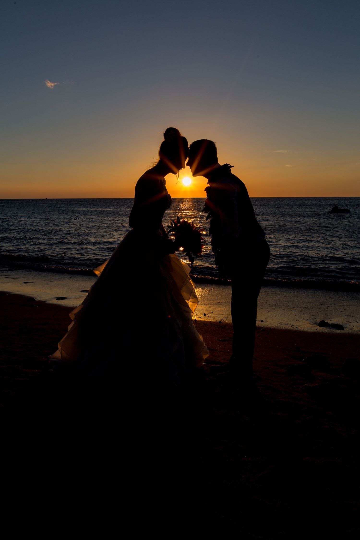 Silhouette of a couple in wedding attire sharing an intimate moment at sunset on a beach with ocean backdrop.
