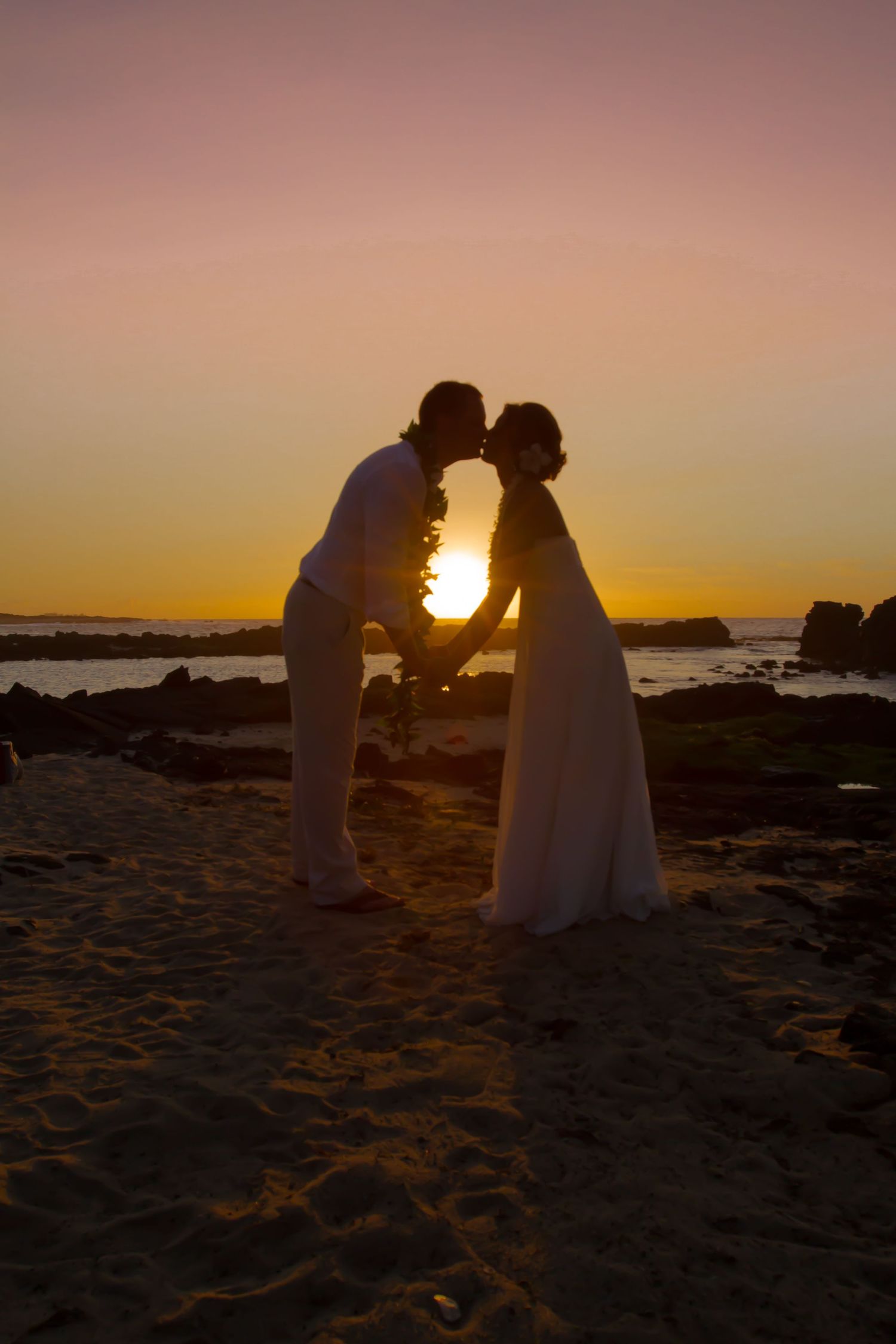 A silhouetted couple in wedding attire share an intimate moment on a beach at sunset with golden light between them in the Kona coast.