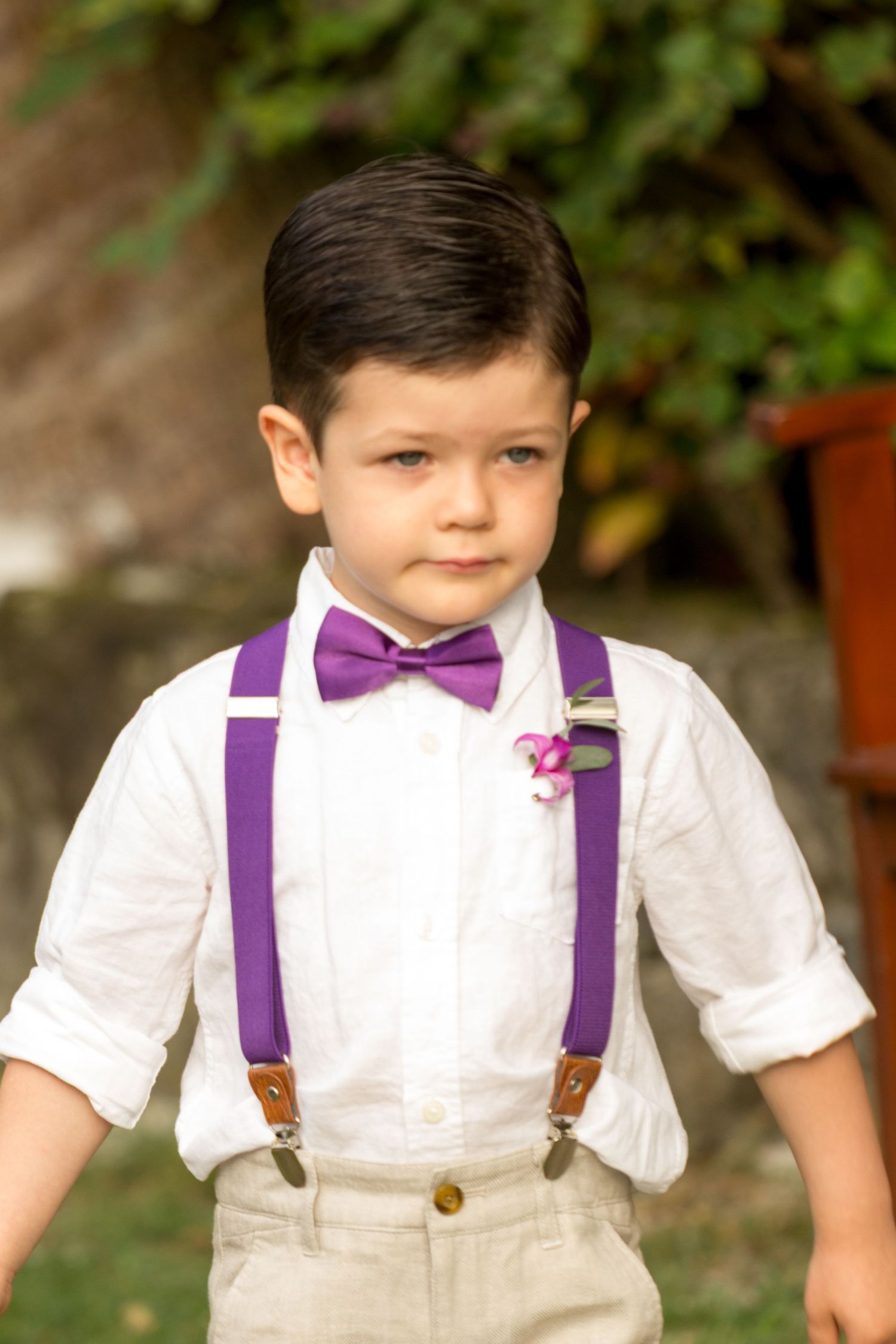 Young ring bearer in white shirt, khaki pants, purple bow tie, and suspenders in a wedding on the lawn of the Outrigger Kona Resort & Spa.