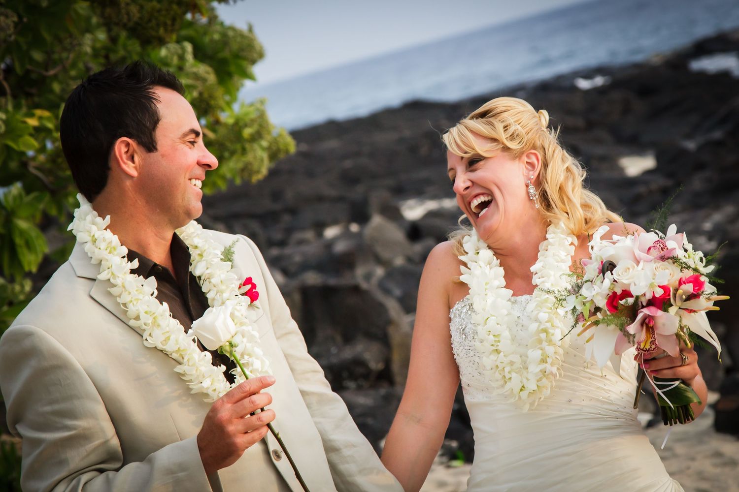 A joyful wedding couple wearing leis laugh together against a rocky Hawaiian coastline backdrop at the Old Airport beach in Kailua Kona.