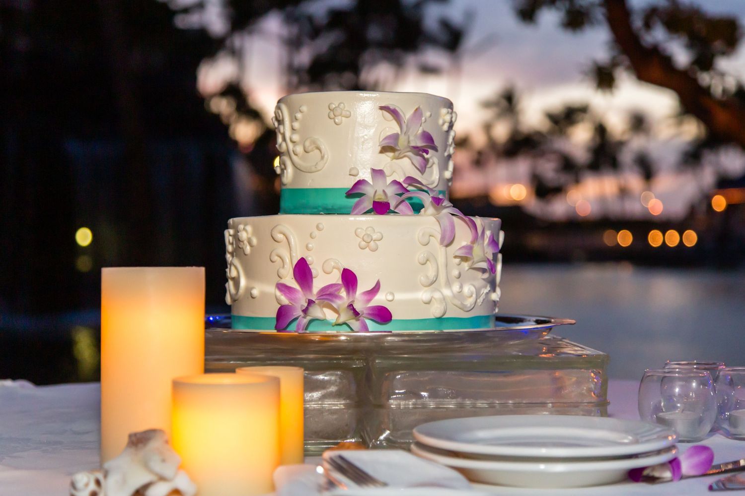 Elegant two-tier wedding cake with purple orchid flowers and glowing candles on a table at sunset.