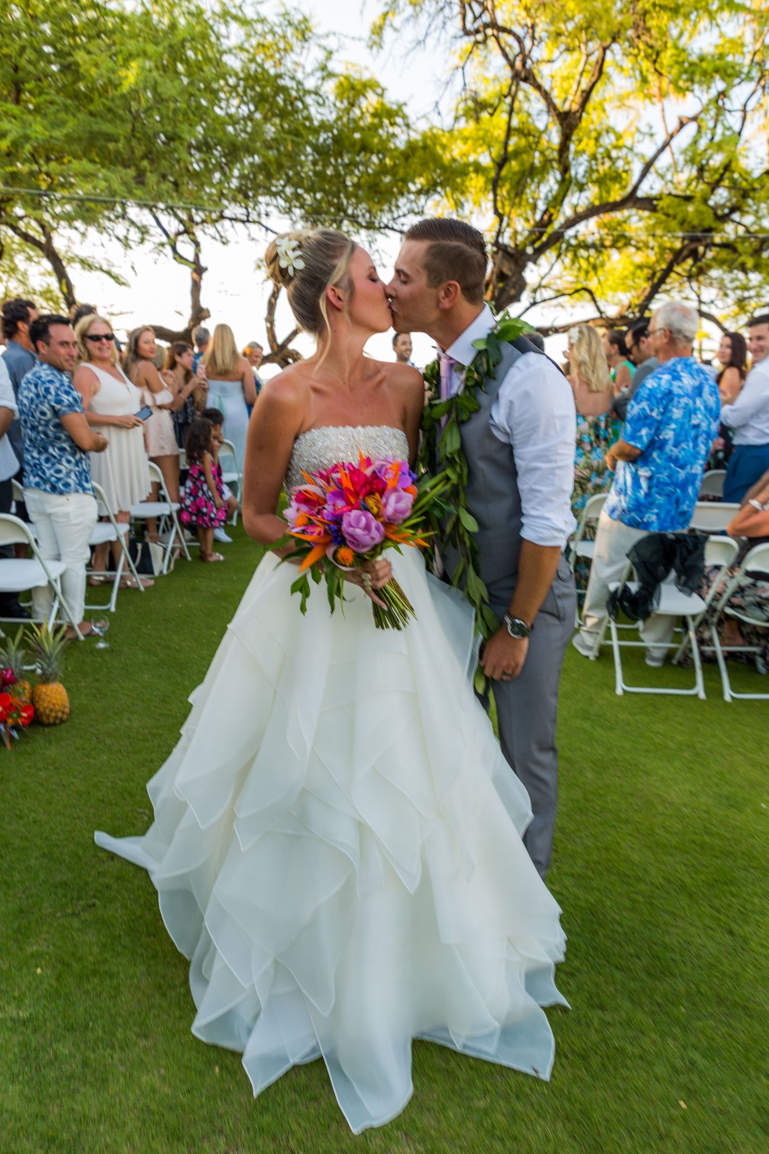 A wedding couple shares their first kiss outdoors on a lawn with guests seated behind them.
