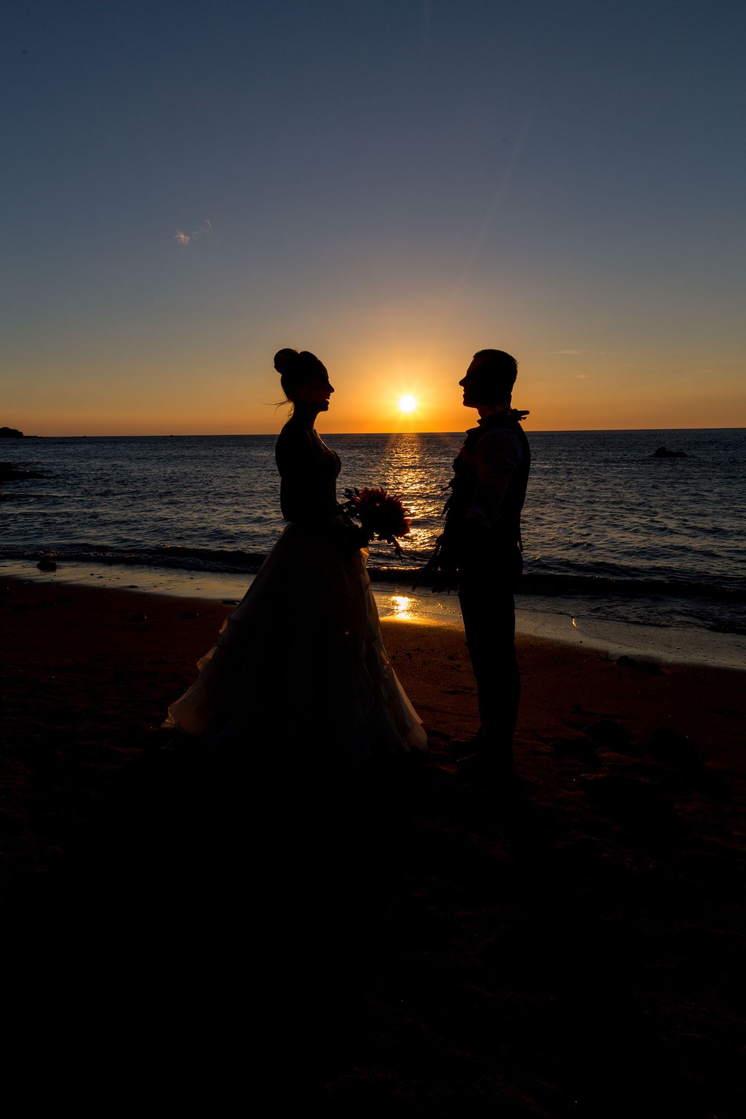 A couple stands in silhouette against the sunset on a beach with golden sunlight reflecting off the ocean waves.