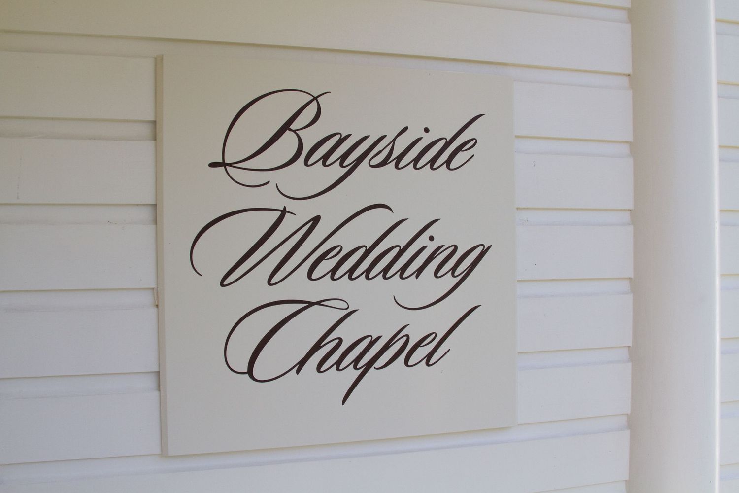 Black artistic script text displaying Bayside wedding chapel on white painted wood paneled wall at the Outrigger hotel in Kona.