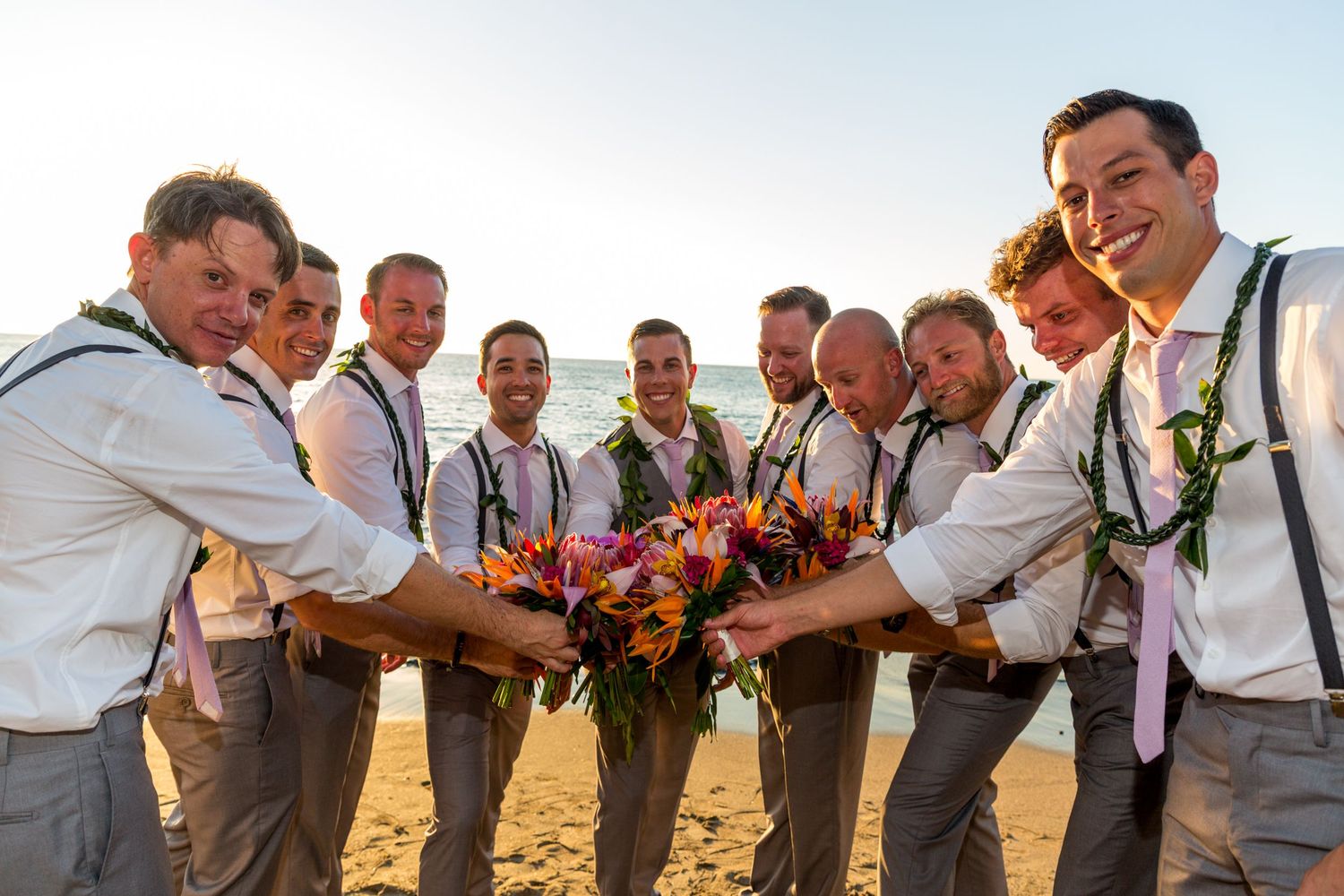 Groomsmen lined up in a row wearing beige vests and suspenders create a playful photoshoot moment at sunset.