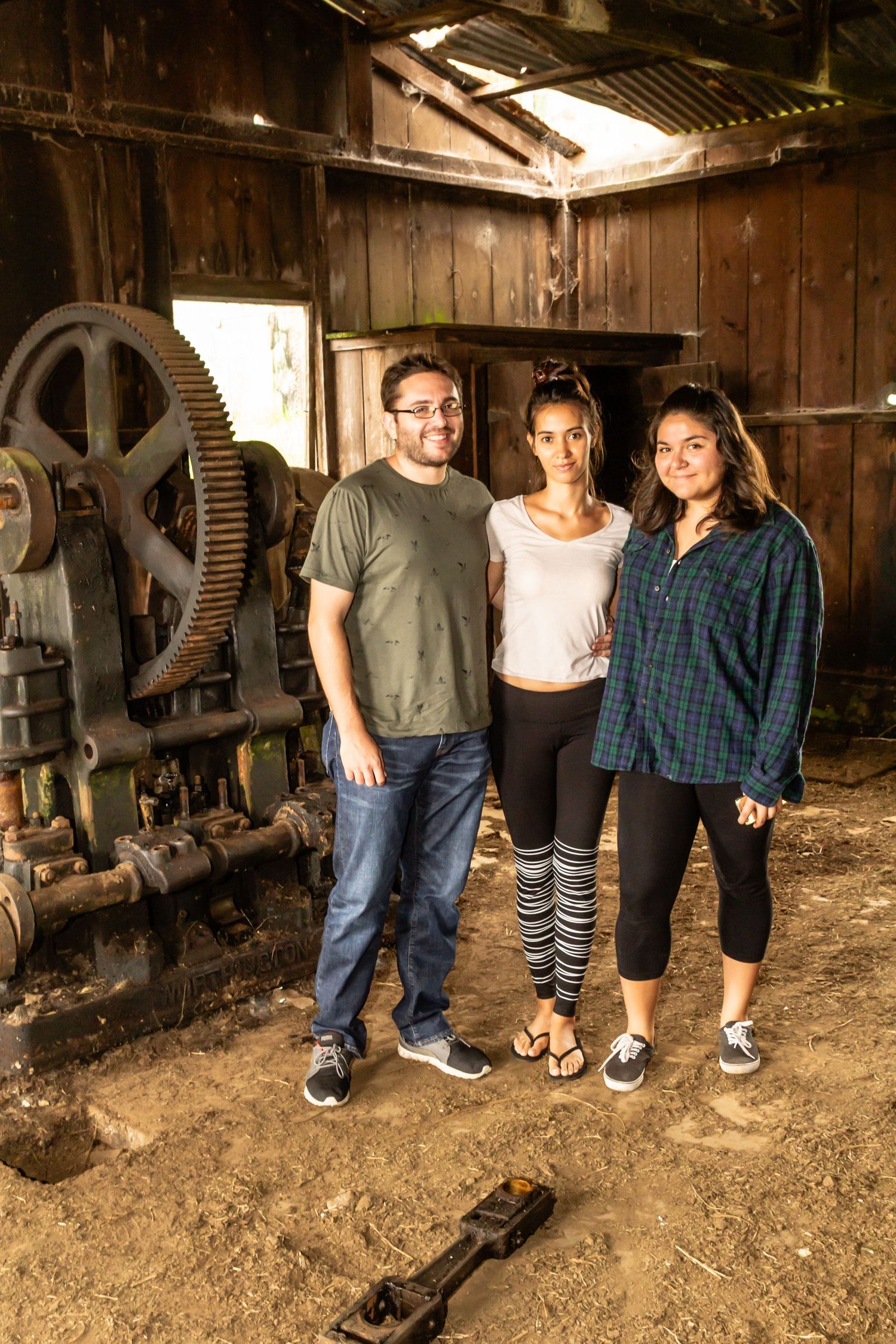 Three people stand together inside a rustic wooden barn with vintage machinery. Photo taken on the Mana Road in Waimea, Big Island of Hawaii