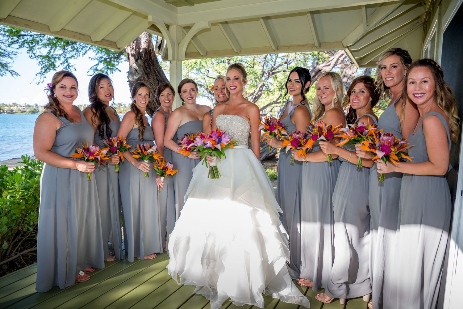 Wedding party in matching light gray bridesmaid dresses holds vibrant fall-colored bouquets at outdoor waterfront gazebo.