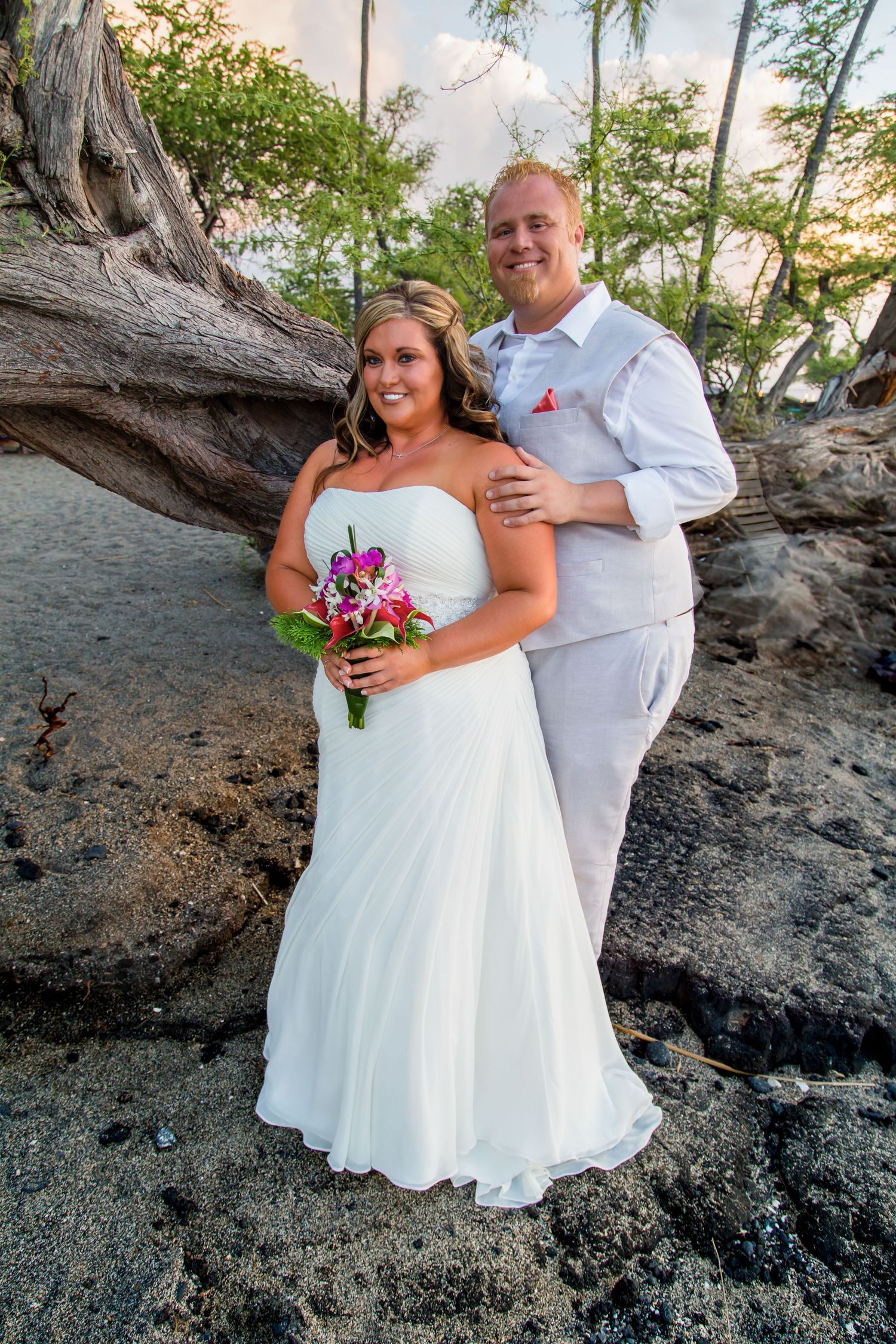 The wedding couple poses at Anaehoomalu Bay beach on the Big Island of Hawaii with the bride holding a pink bouquet.
