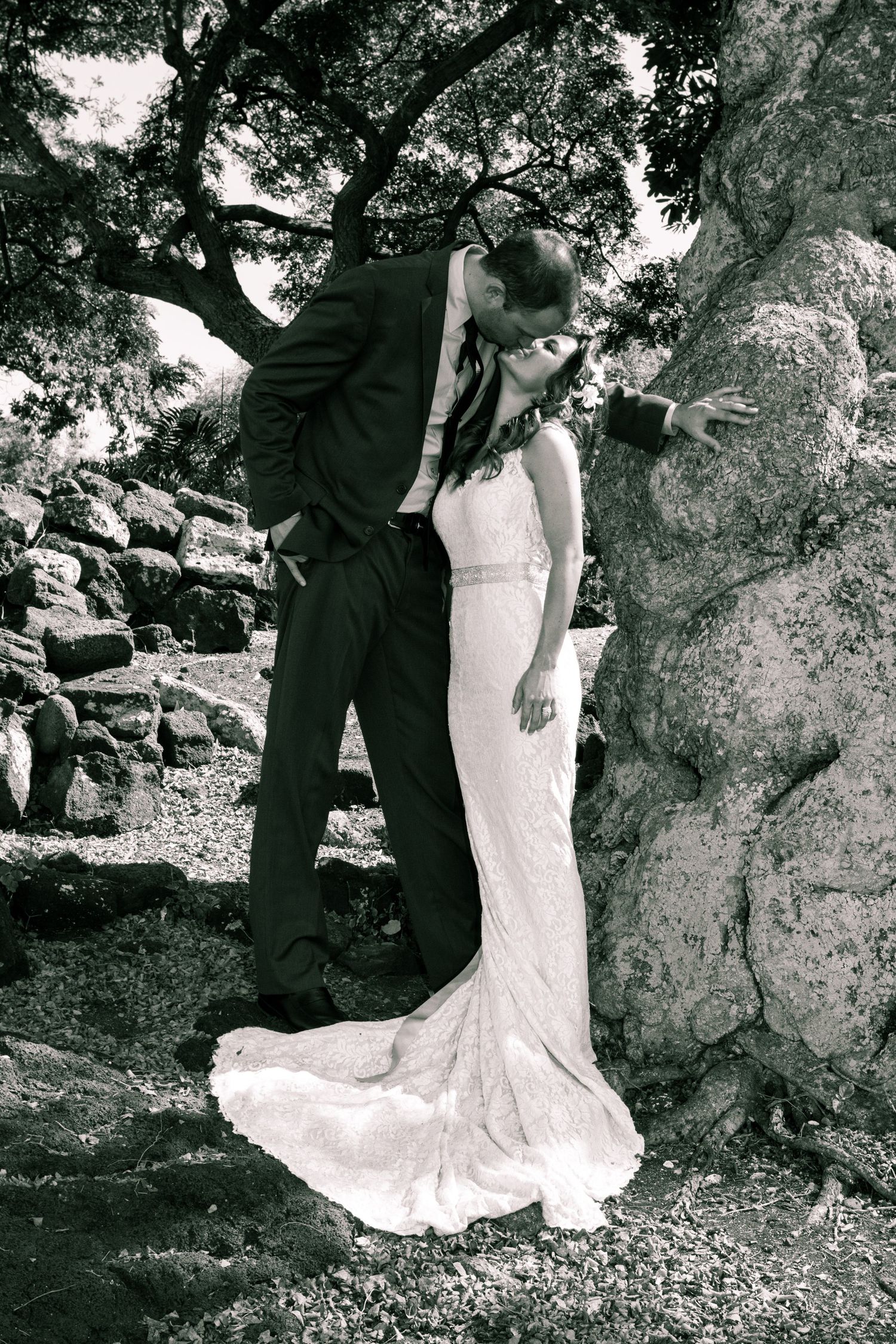 A bride in a fitted lace gown and groom in dark suit share a romantic moment against a rocky natural backdrop.