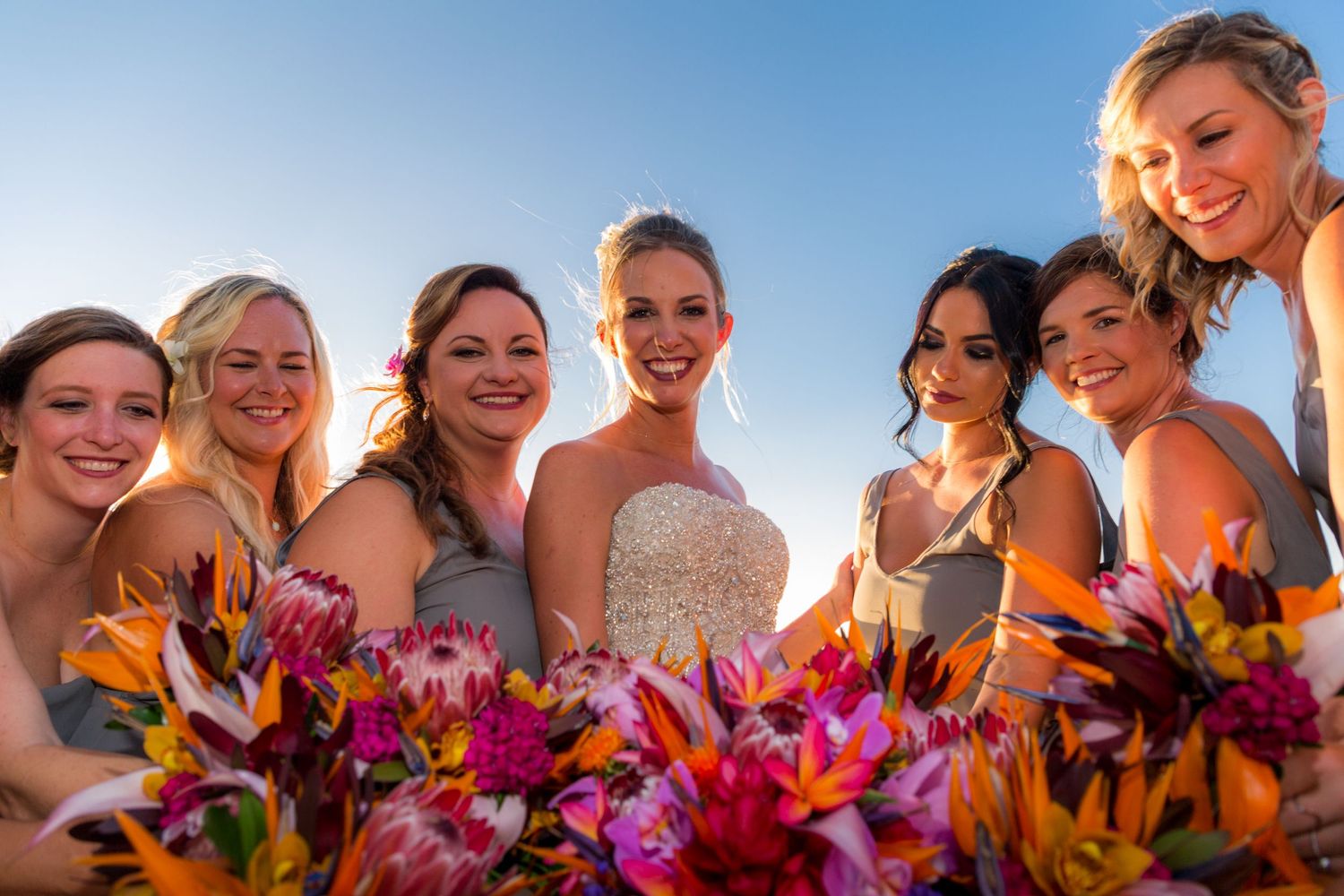 A bridal party holding vibrant orange and pink flower bouquets poses together against a bright blue sky at sunset.