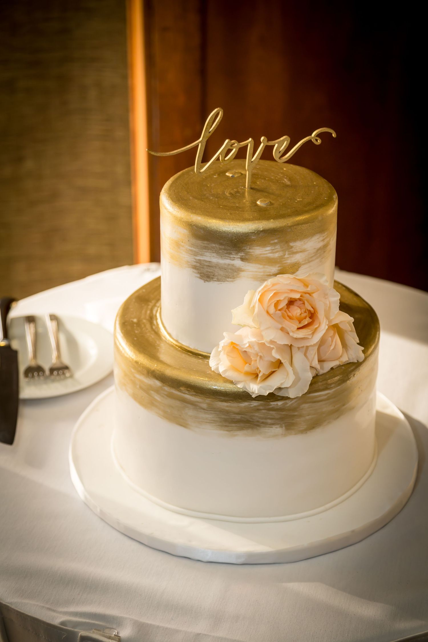 Two-tier white and gold wedding cake decorated with peach roses and 'love' cake topper.