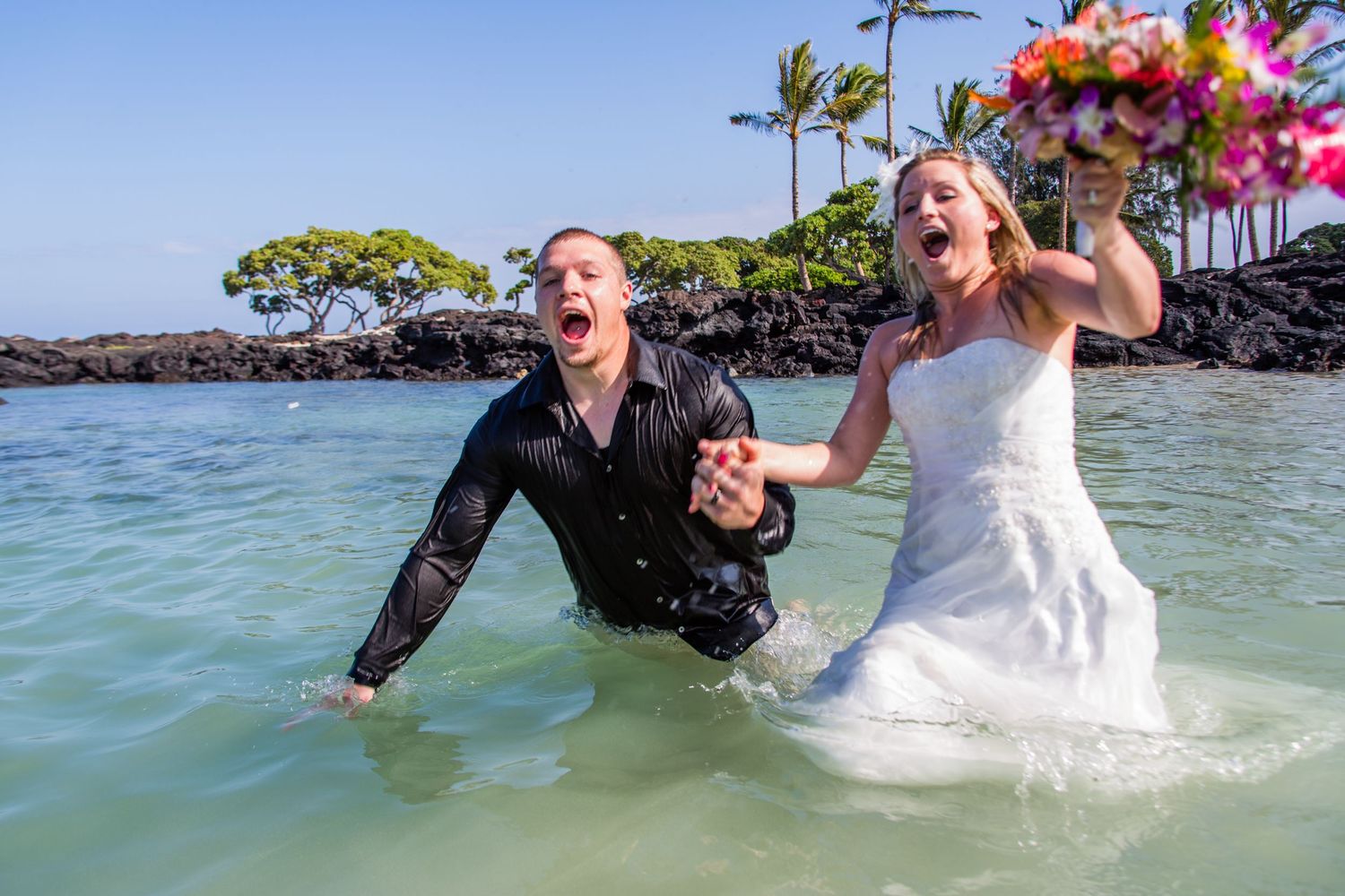 Newlyweds playfully splash in crystal clear tropical waters near palm trees and colorful flowers at Kikaua Beach in Kailua Kona, Hawaii.