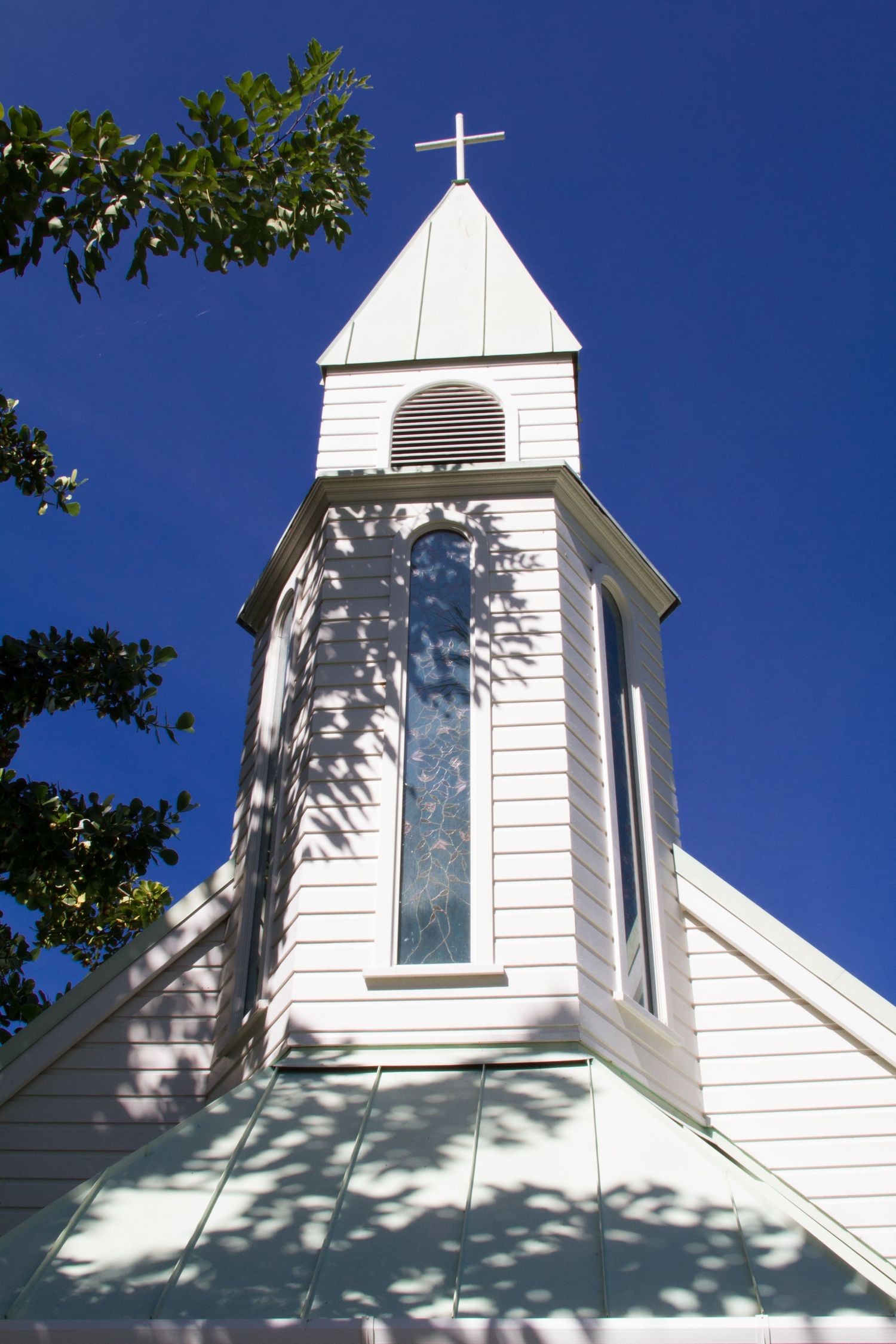 White wooden church steeple with tall window and cross rises against bright blue sky with green leaves framing the corner.