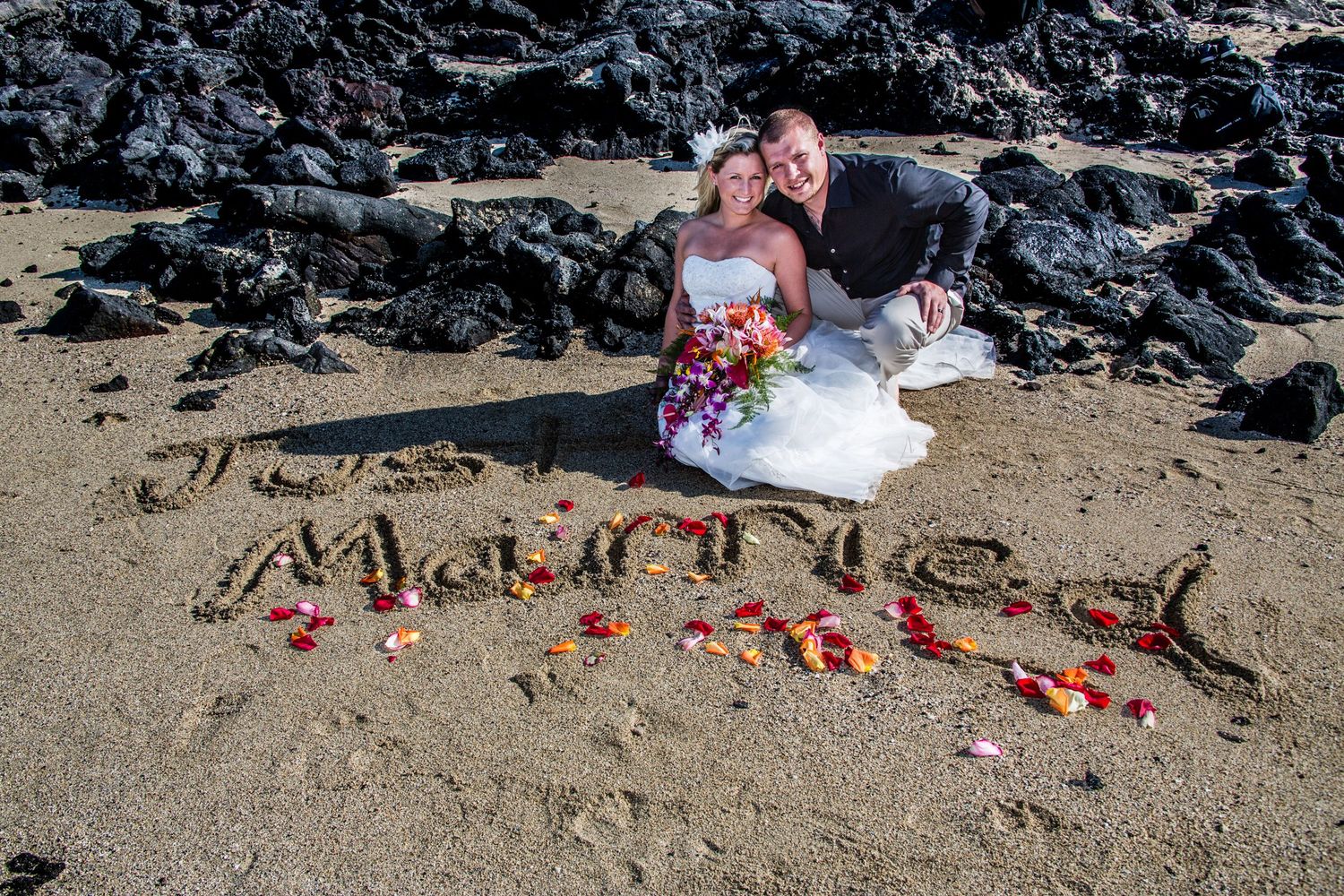 A couple sit on volcanic beach rocks beside 'Married' written in sand with tropical flowers in Hawaii at Kikaua beach in Kailua Kona, Hawaii