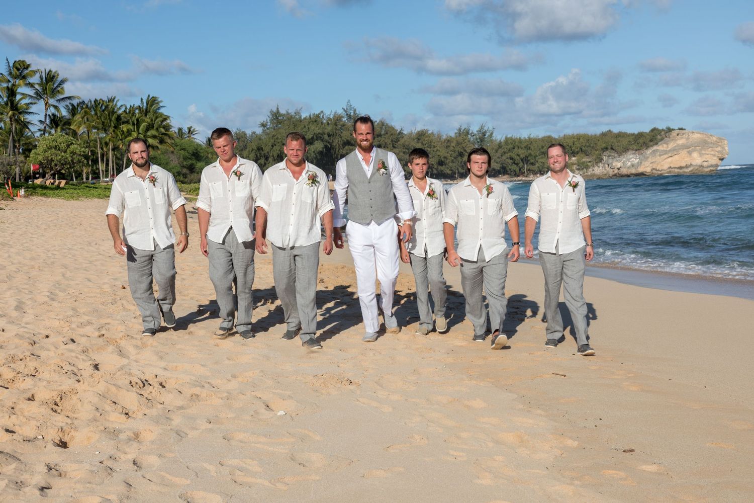 Groomsmen in white shirts and gray pants posing together on a sandy beach with cliffs in the background in Kauai, Hawaii.