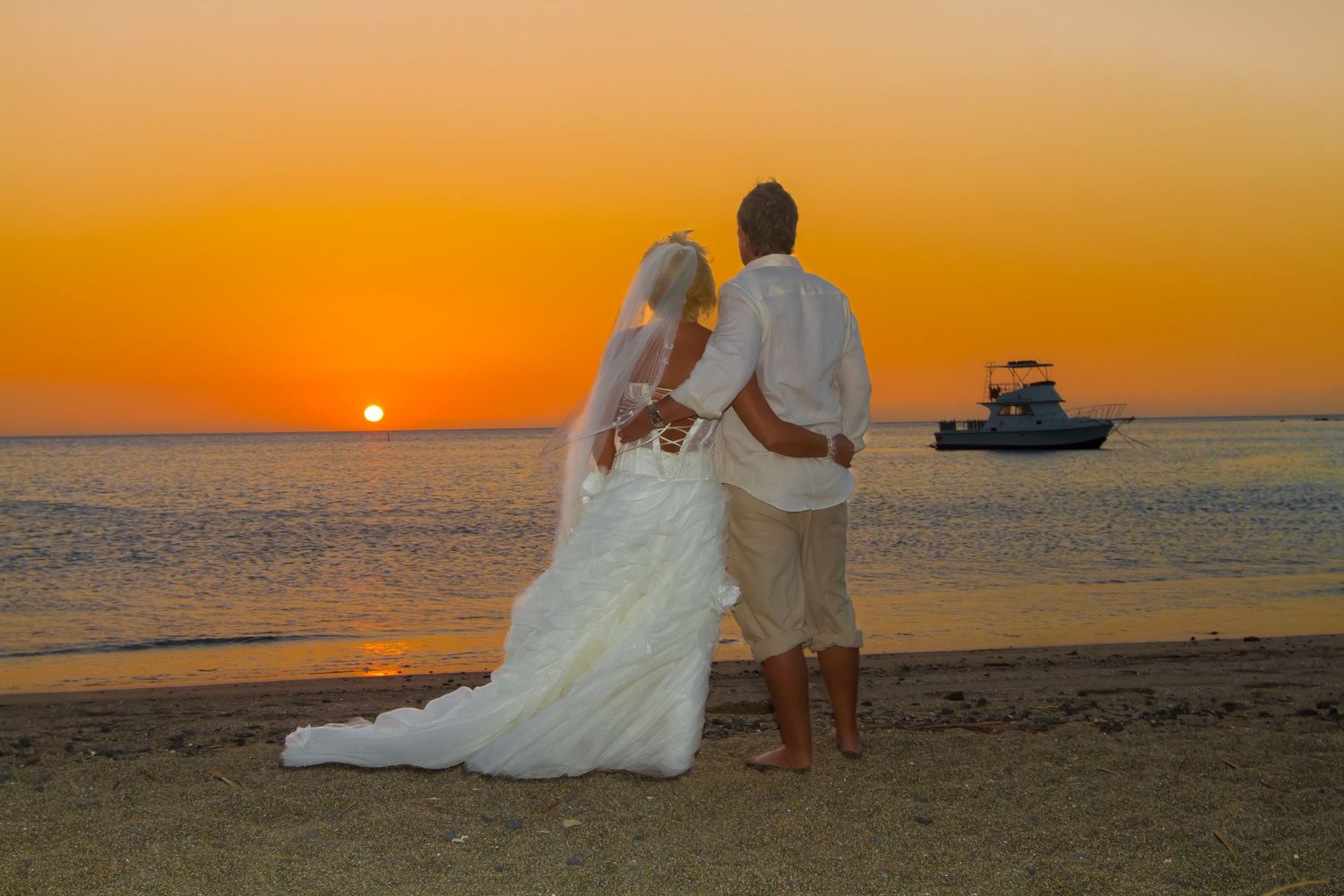 A couple in wedding attire watches the sunset over the ocean with a boat silhouetted on the horizon.