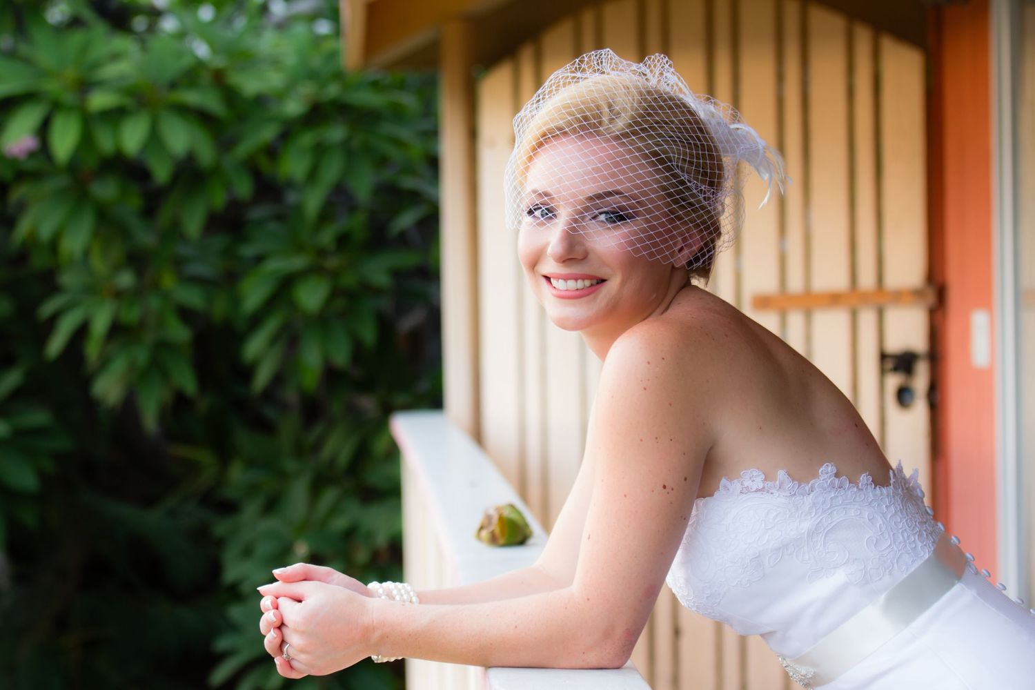A radiant bride in a strapless white gown leans on a wooden railing with a joyful smile before a wedding at a private state in Kailua Kona.