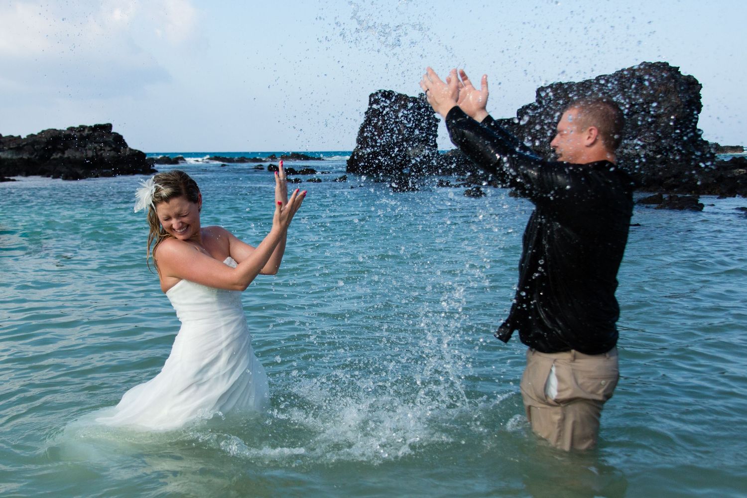 Newlyweds splash crystal clear turquoise ocean waters near dark rocky formations during a trash the dress photoshoot at Kikaua beach in Kona