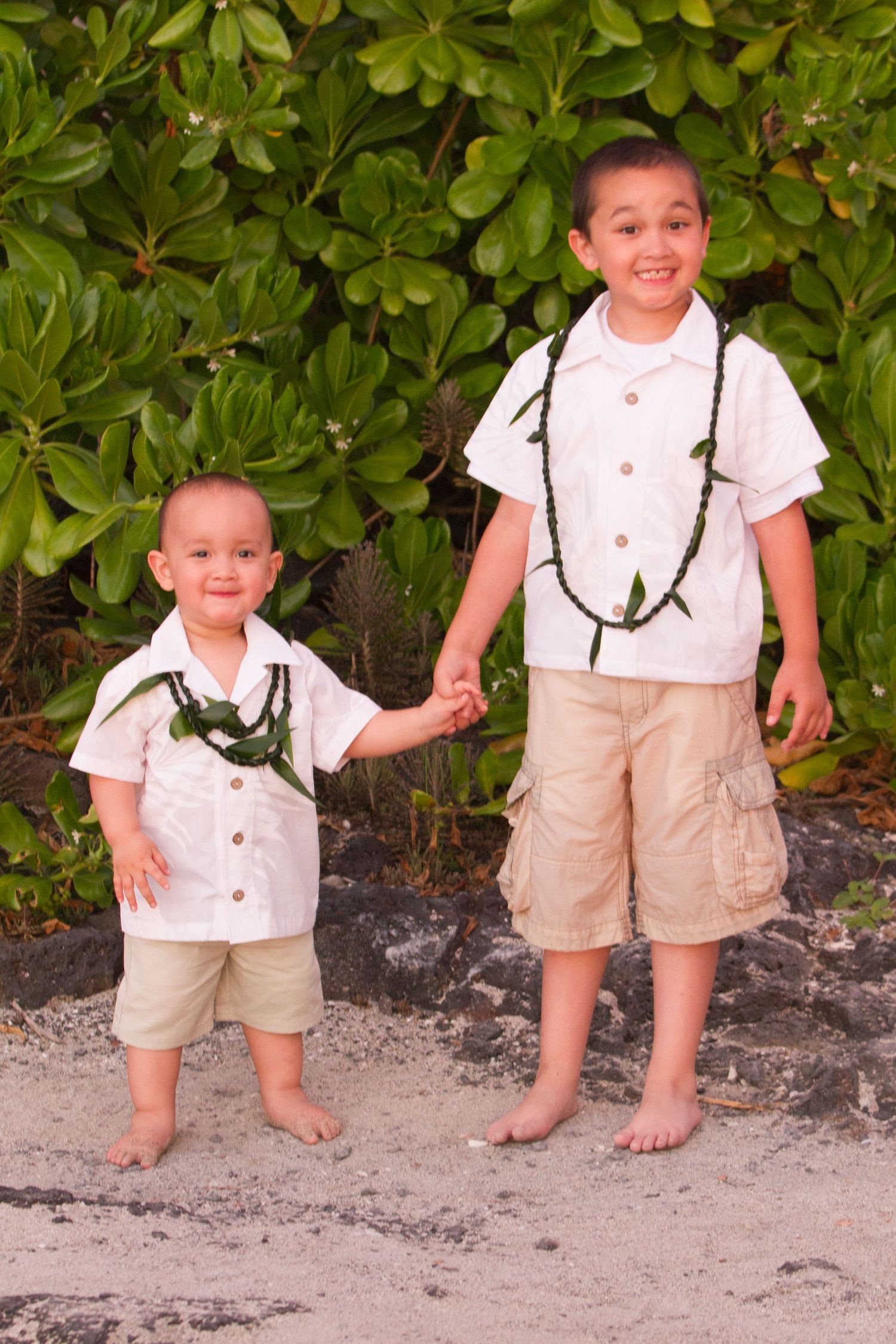 Two children in matching white shirts and khaki shorts standing on beach with tropical foliage at Konokohau beach in Kailua Kona.