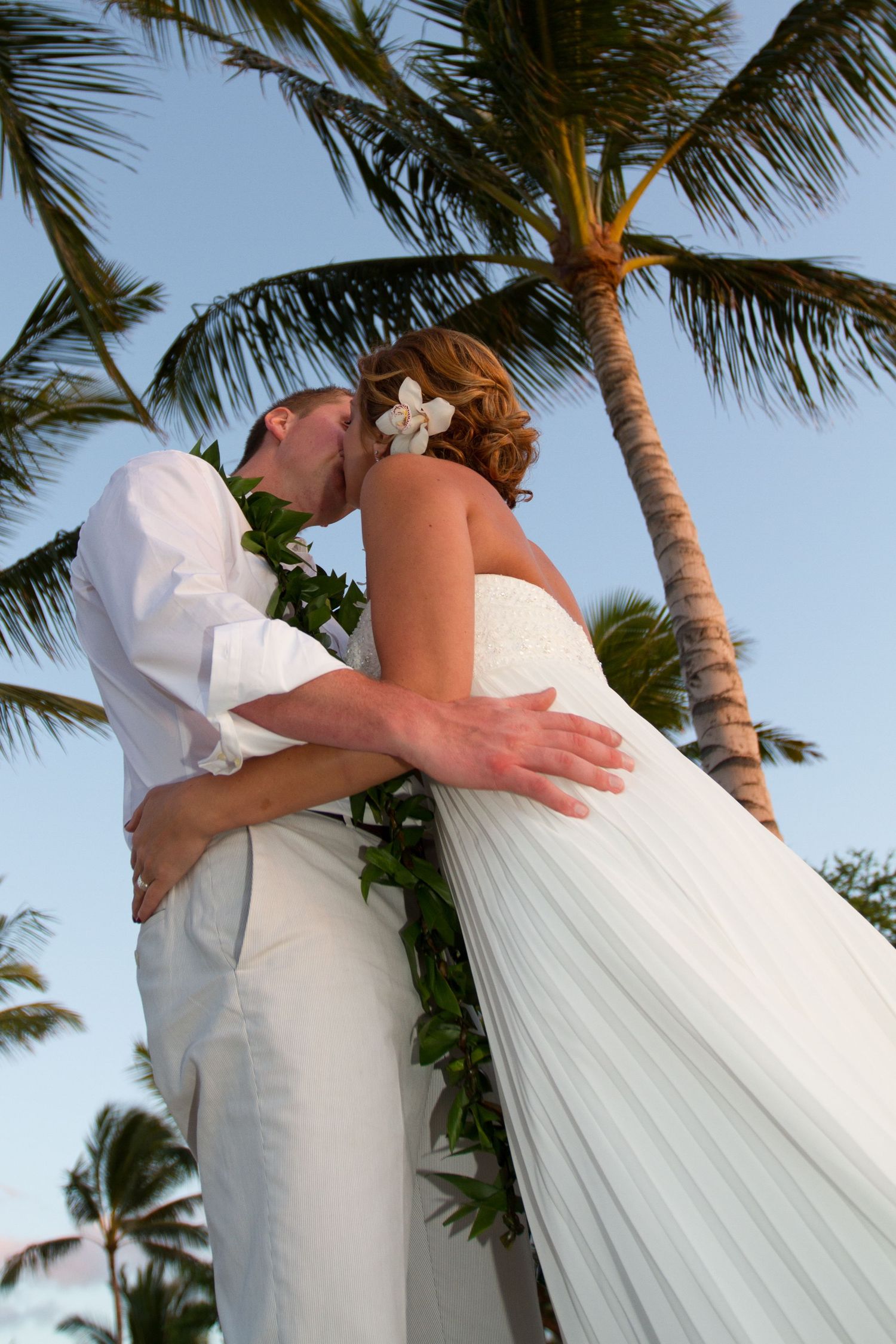 A romantic wedding couple embraces under tall palm trees on a tropical Hawaii beach at sunset.