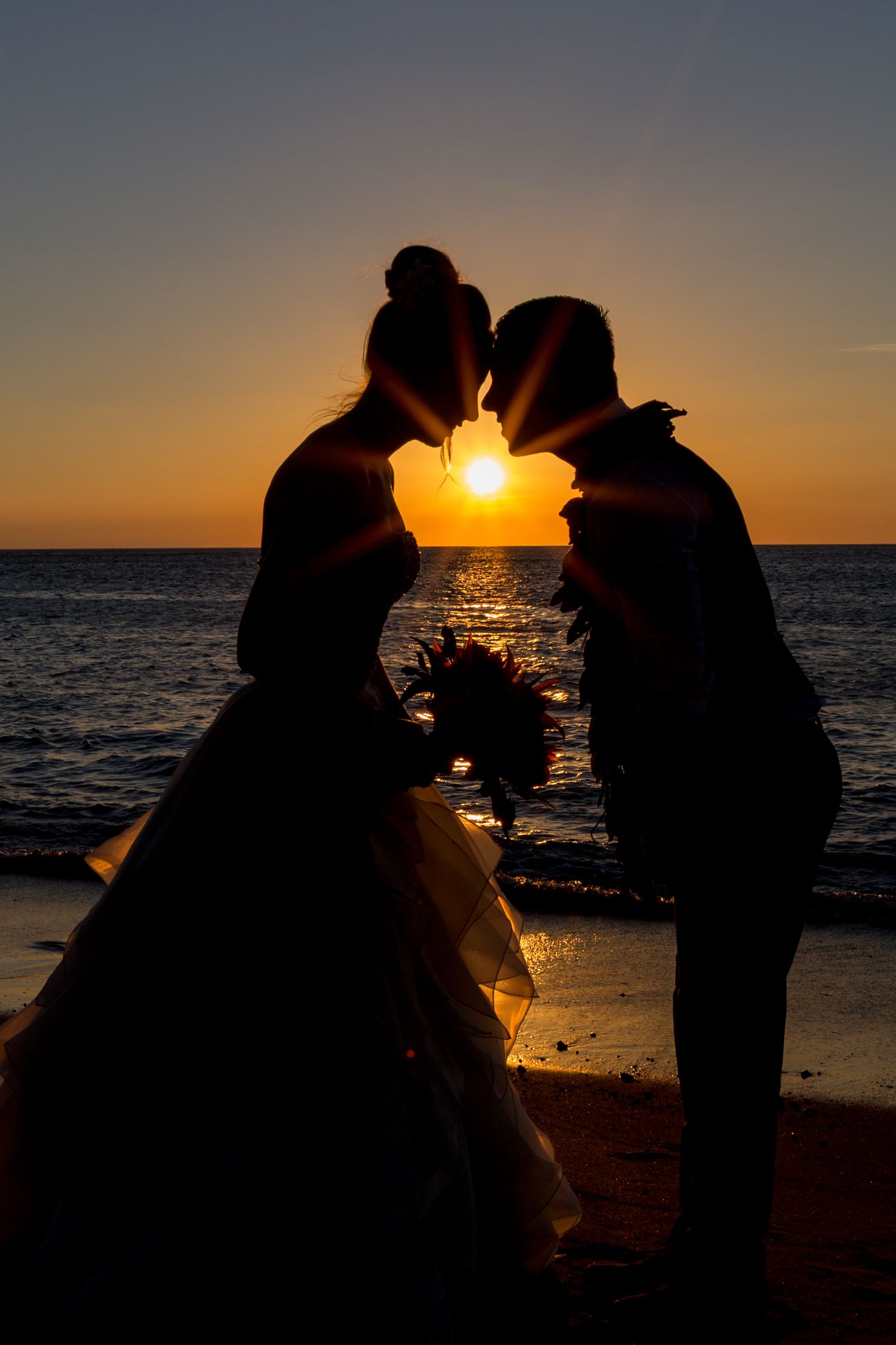 Silhouetted couple shares romantic moment on beach at sunset with ocean waves and glowing sun in background.