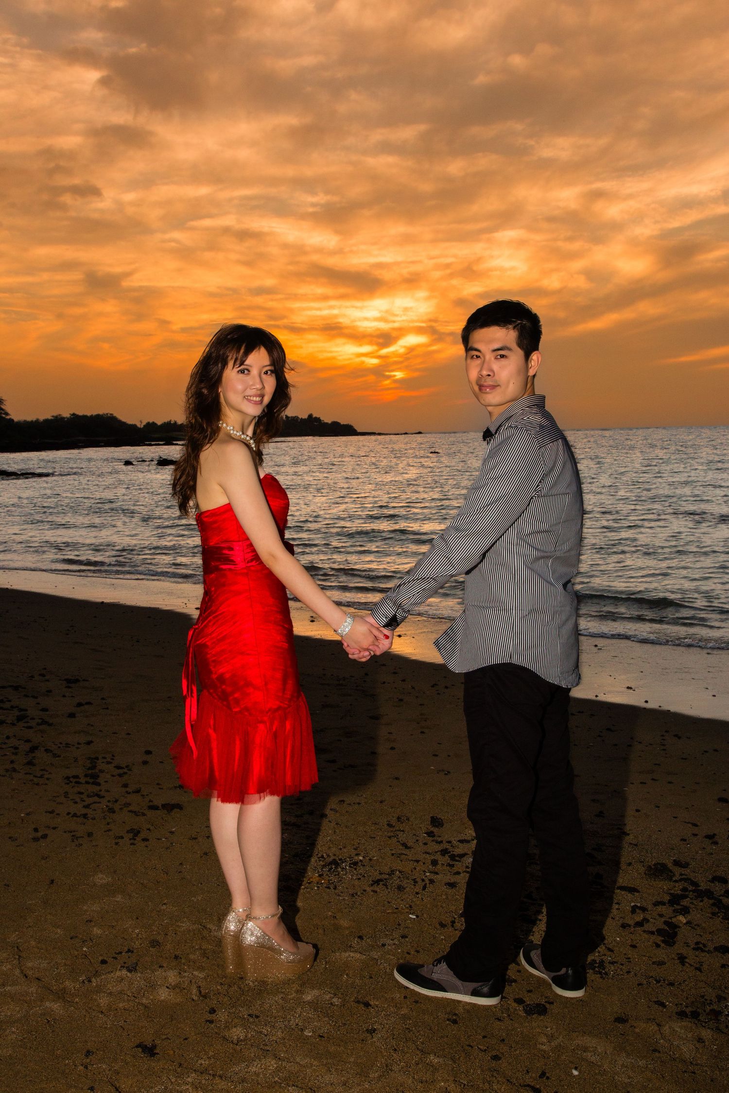 Couple poses for engagement photo at sunset beach, woman in red dress holding hands with partner at Anaehoomalu Bay beach near Kailua Kona.