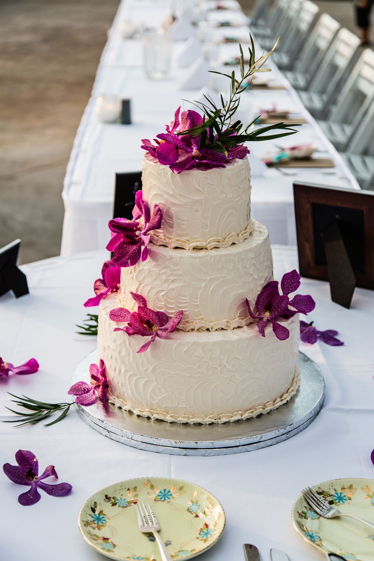 Three-tiered white wedding cake decorated with purple orchids and greenery on a white tablecloth.