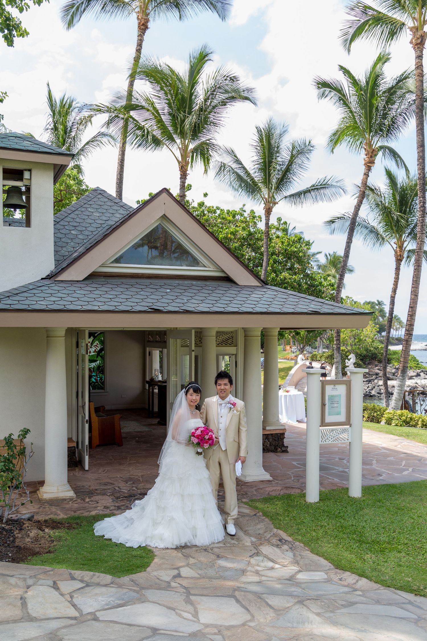 Newlyweds pose under palm trees outside tropical wedding chapel with stone pathway.