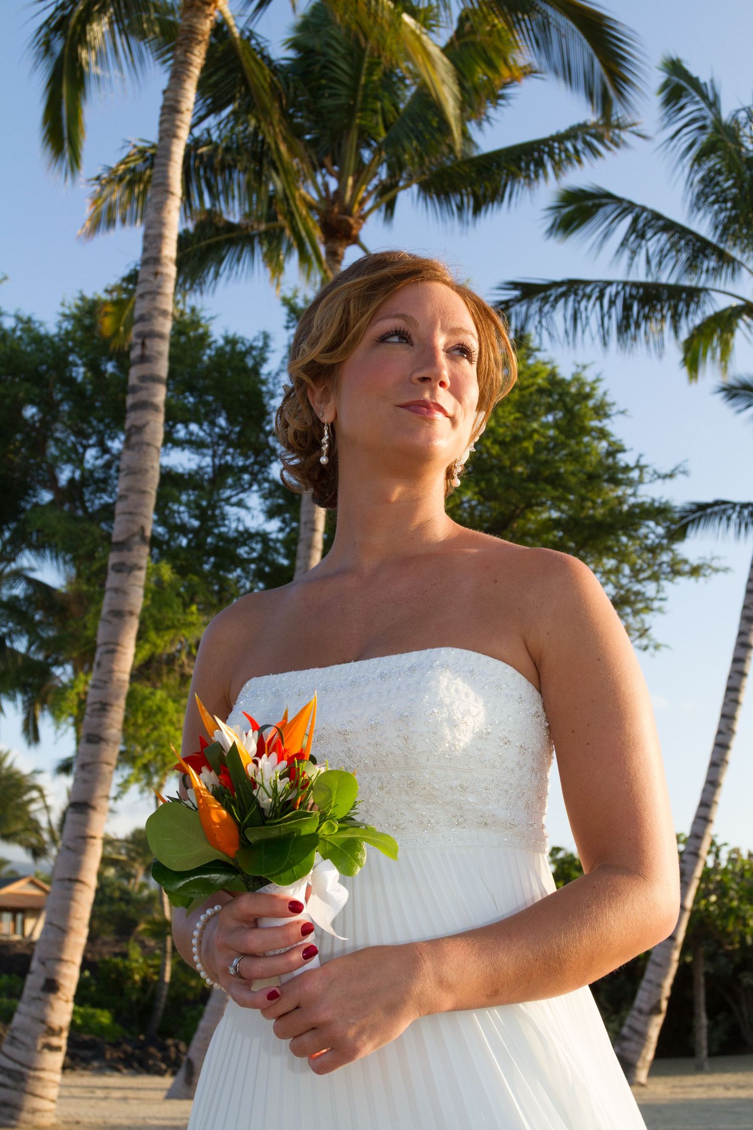 A bride in a white strapless dress holds a tropical bouquet while standing among swaying palm trees on a beach in Kona.