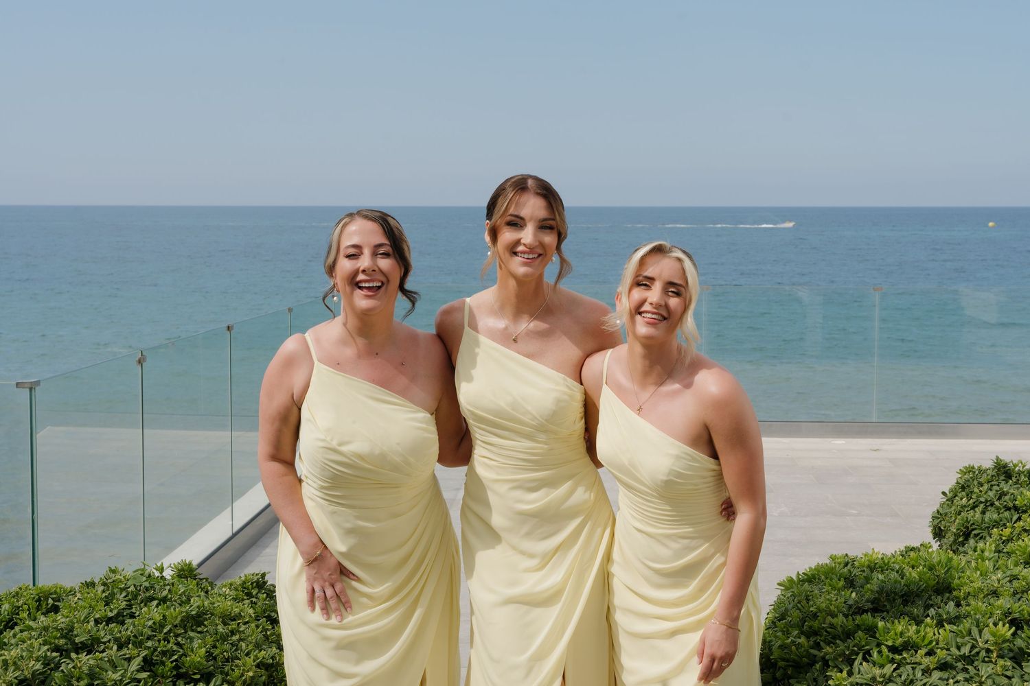 Three bridesmaids in pale yellow dresses smiling by the ocean.