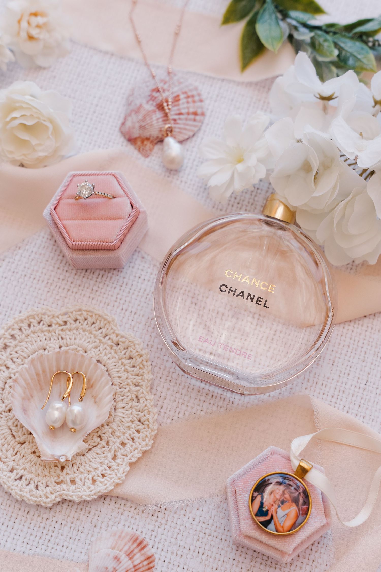 Elegant wedding details featuring Chanel perfume, ring box, seashells and white roses on lace fabric.
