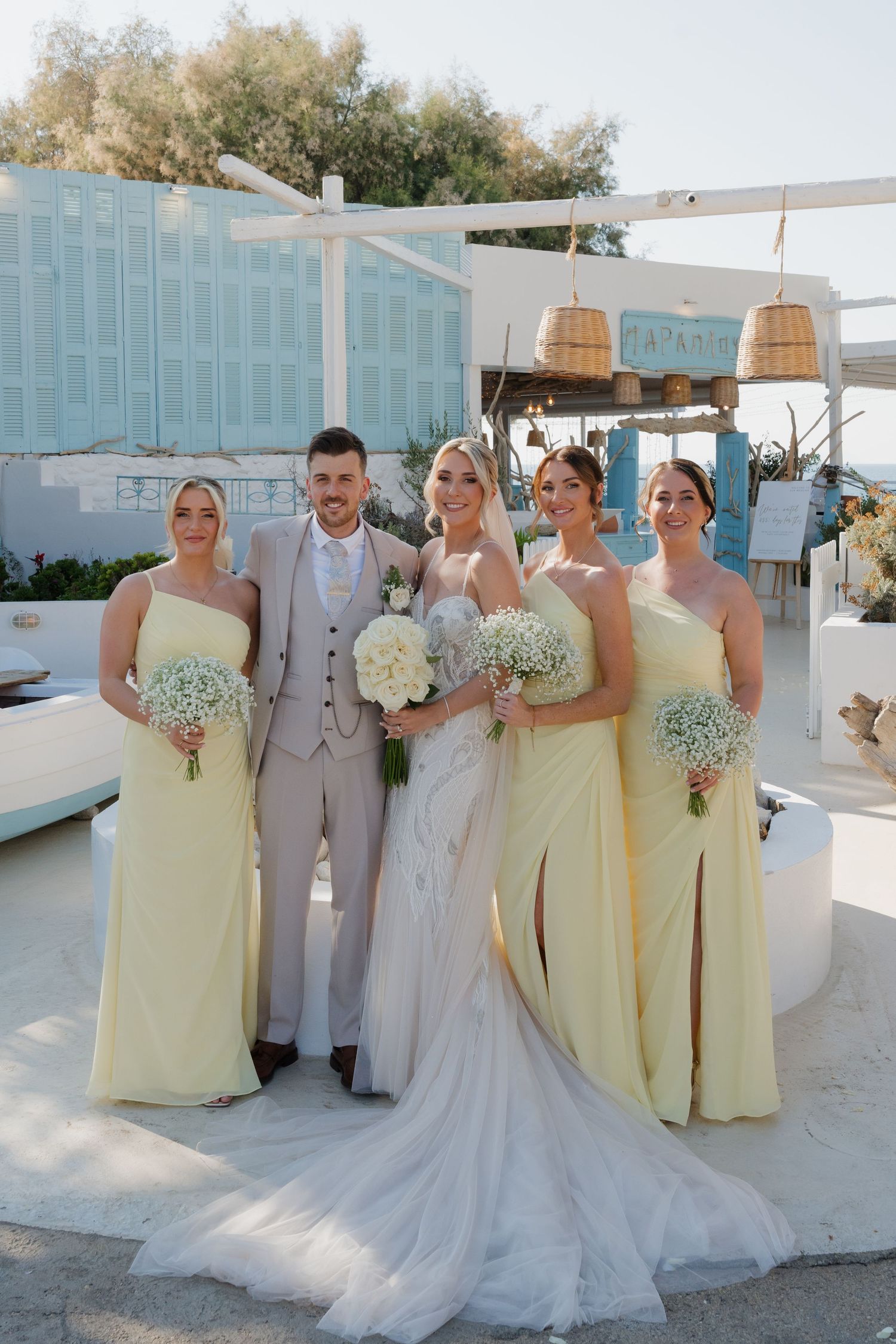 Wedding party in pale yellow bridesmaid dresses and light suits posing against white Mediterranean backdrop.