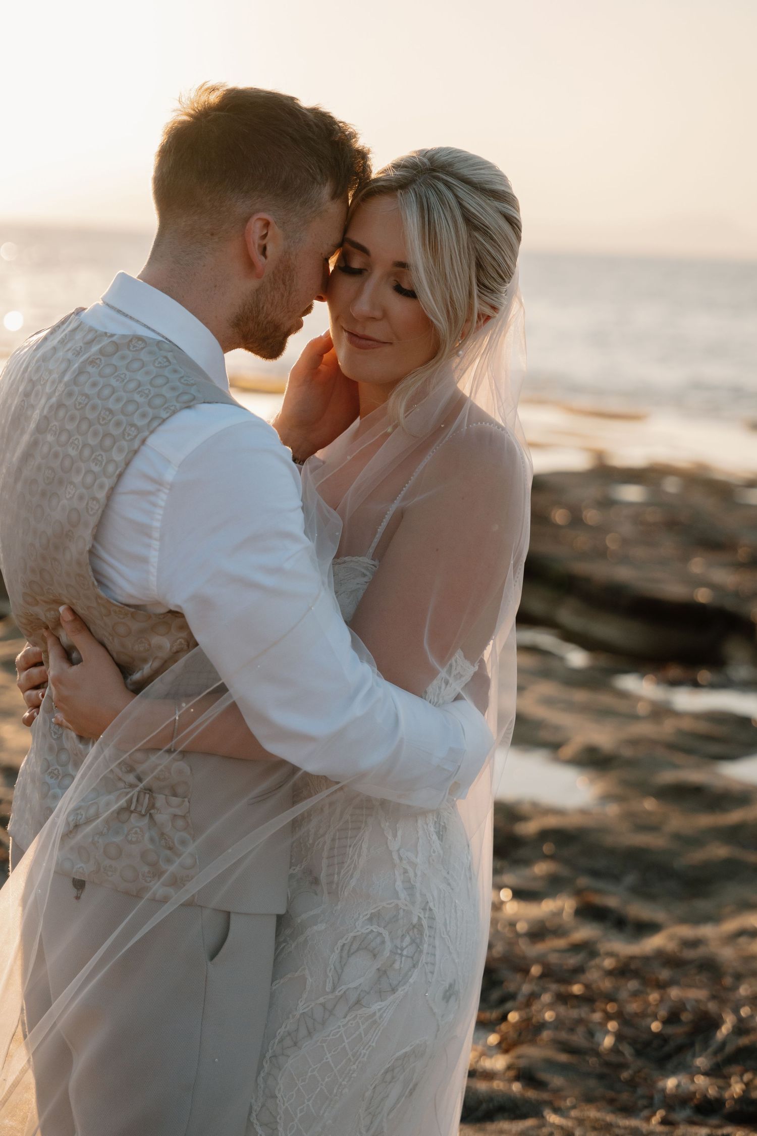Intimate sunset beach portrait of newlyweds embracing against rocky shoreline.