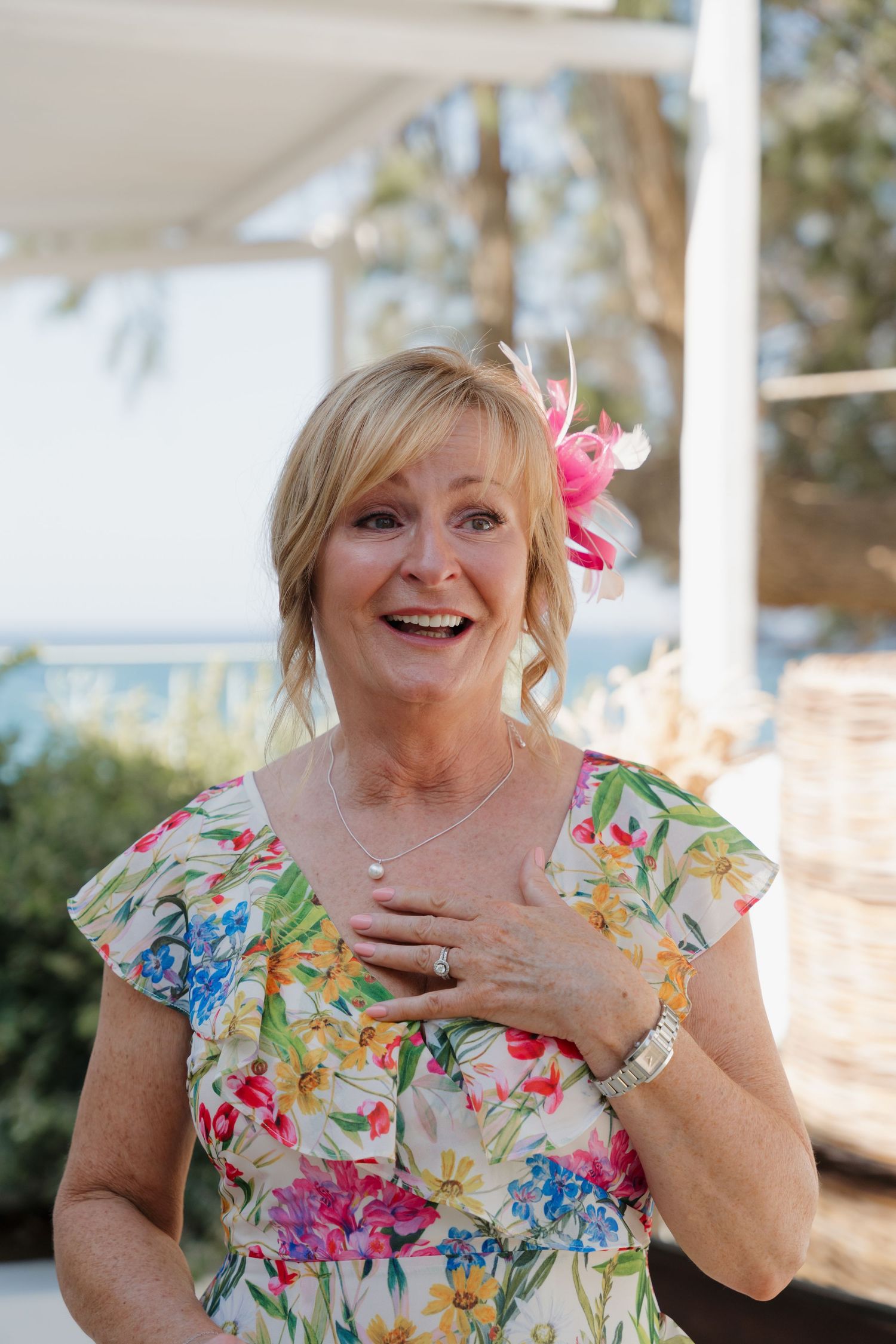 A mature woman in a vibrant floral dress smiles joyfully at a seaside celebration.