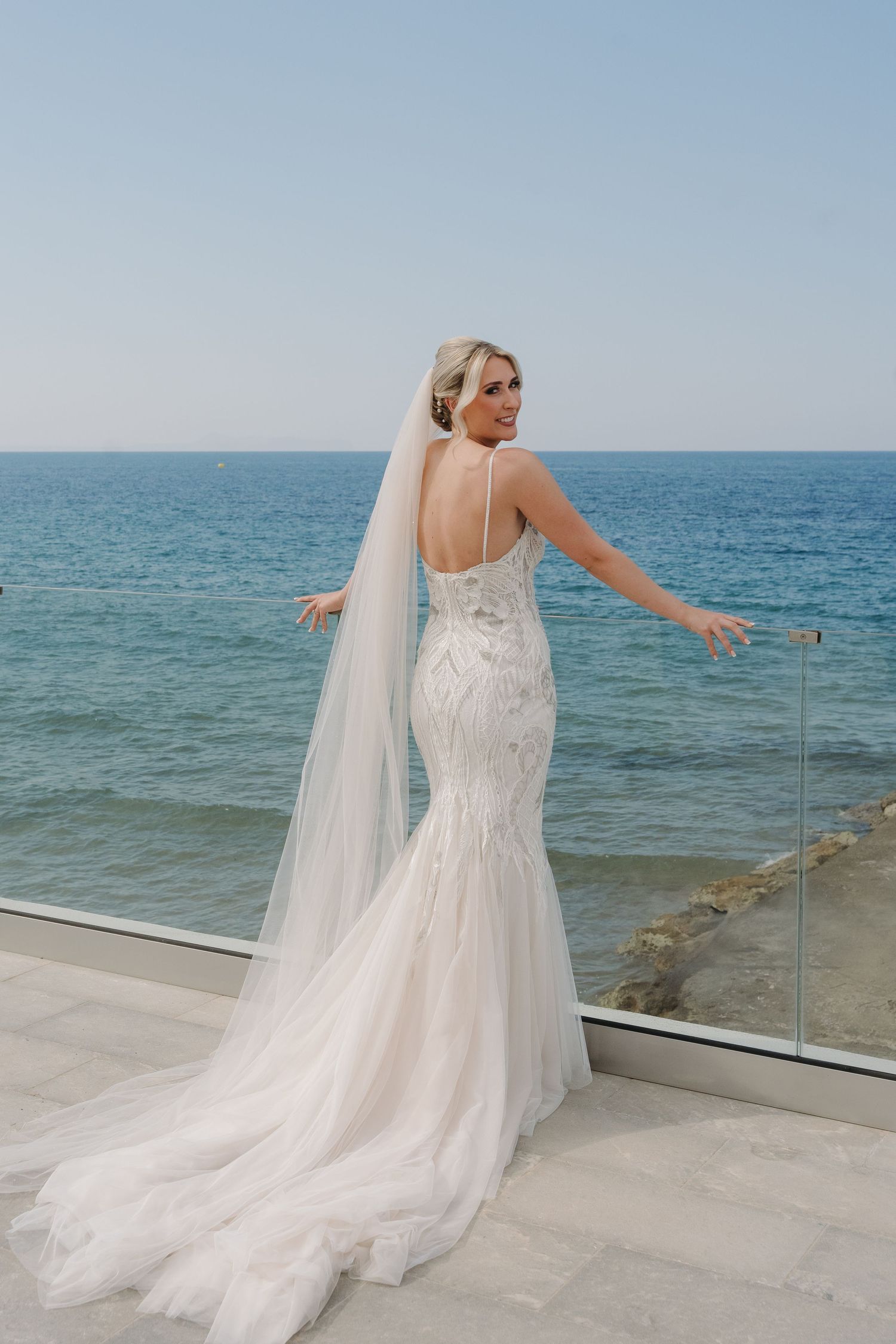 Elegant mermaid wedding dress with long veil photographed against ocean backdrop on seaside terrace.