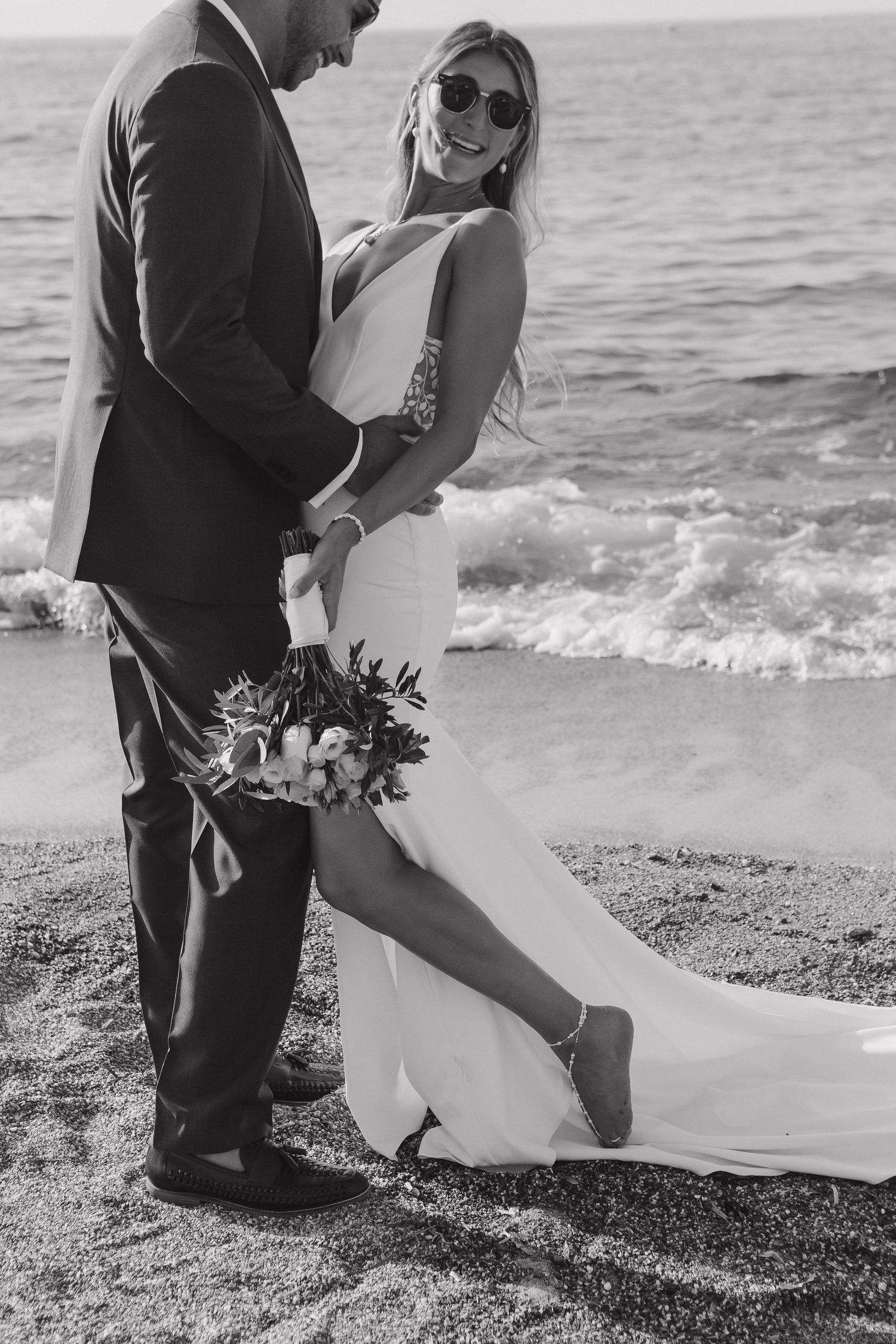 A romantic black and white beach wedding photo featuring a couple embracing by the ocean with a floral bouquet.