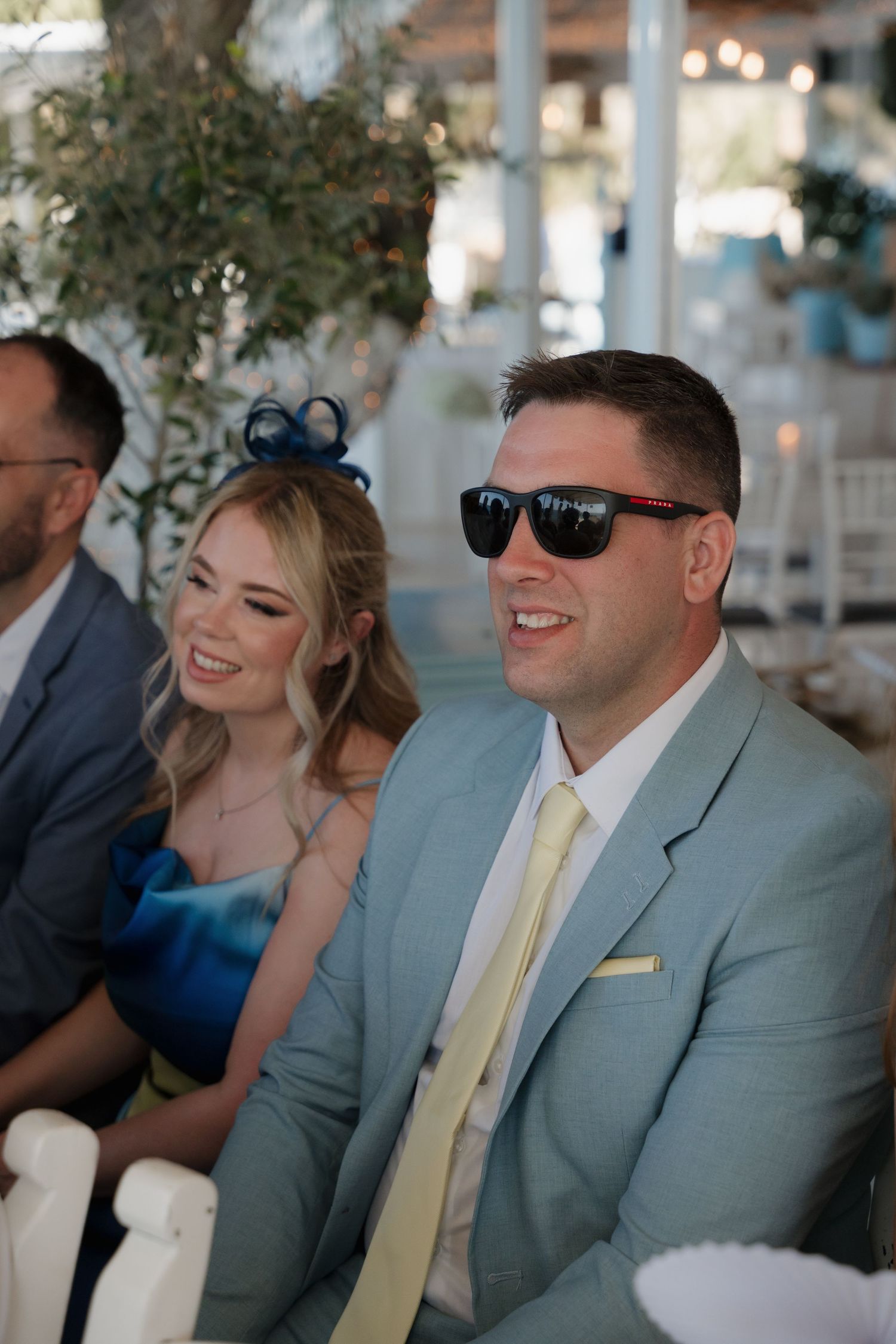 Wedding guests in formal attire seated at outdoor reception with string lights and greenery decor.