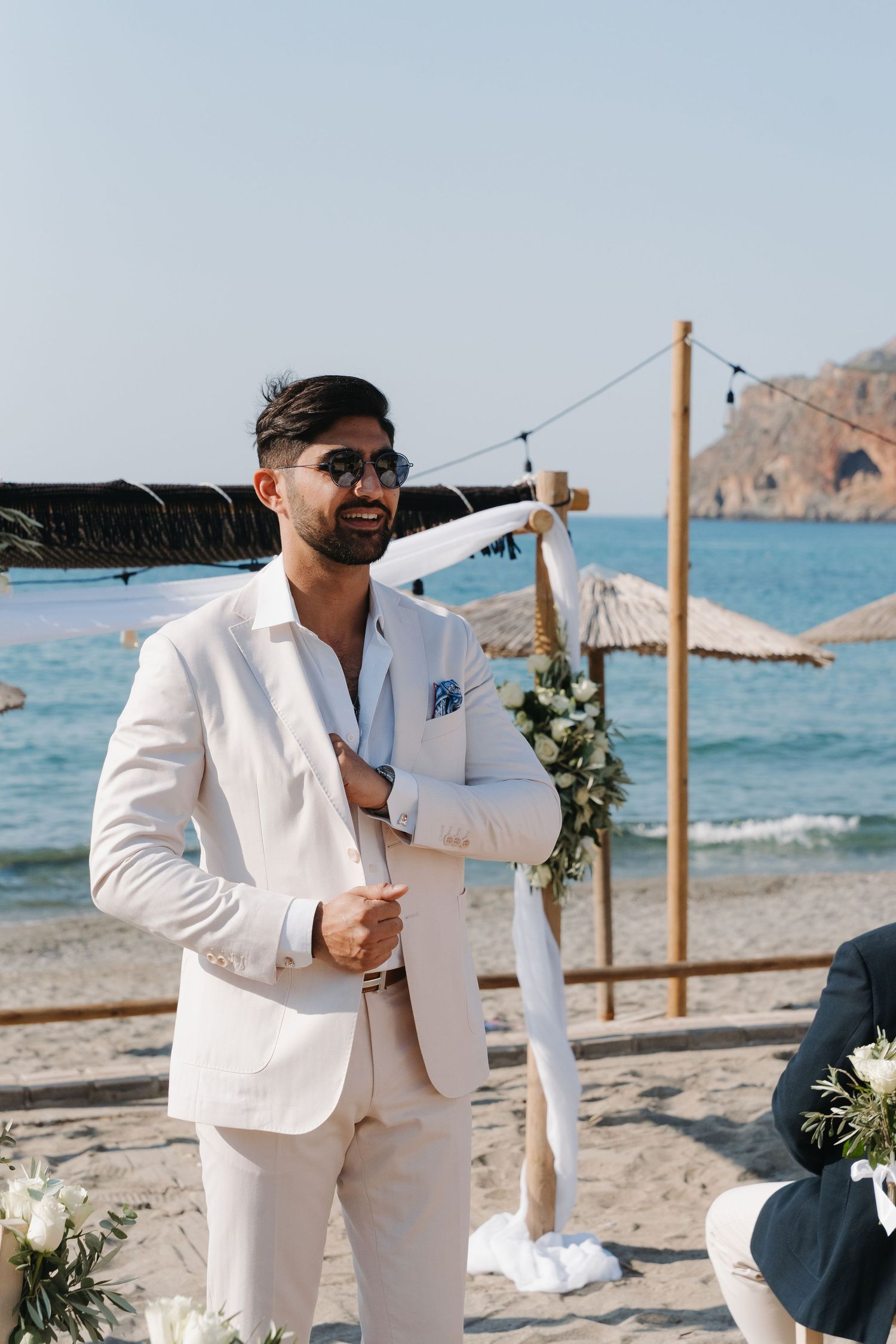 Groomsmen in white suits celebrate on a scenic beach with floral decorations and wooden posts by the ocean.