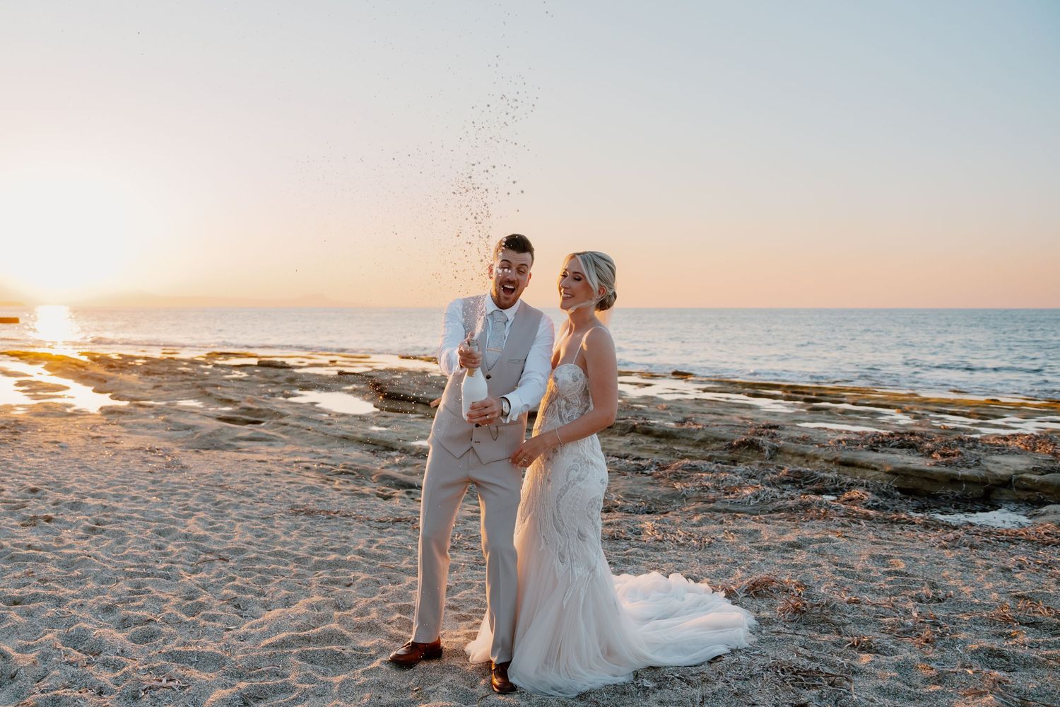 Couple celebrating with champagne on beach at sunset with ocean waves in background.