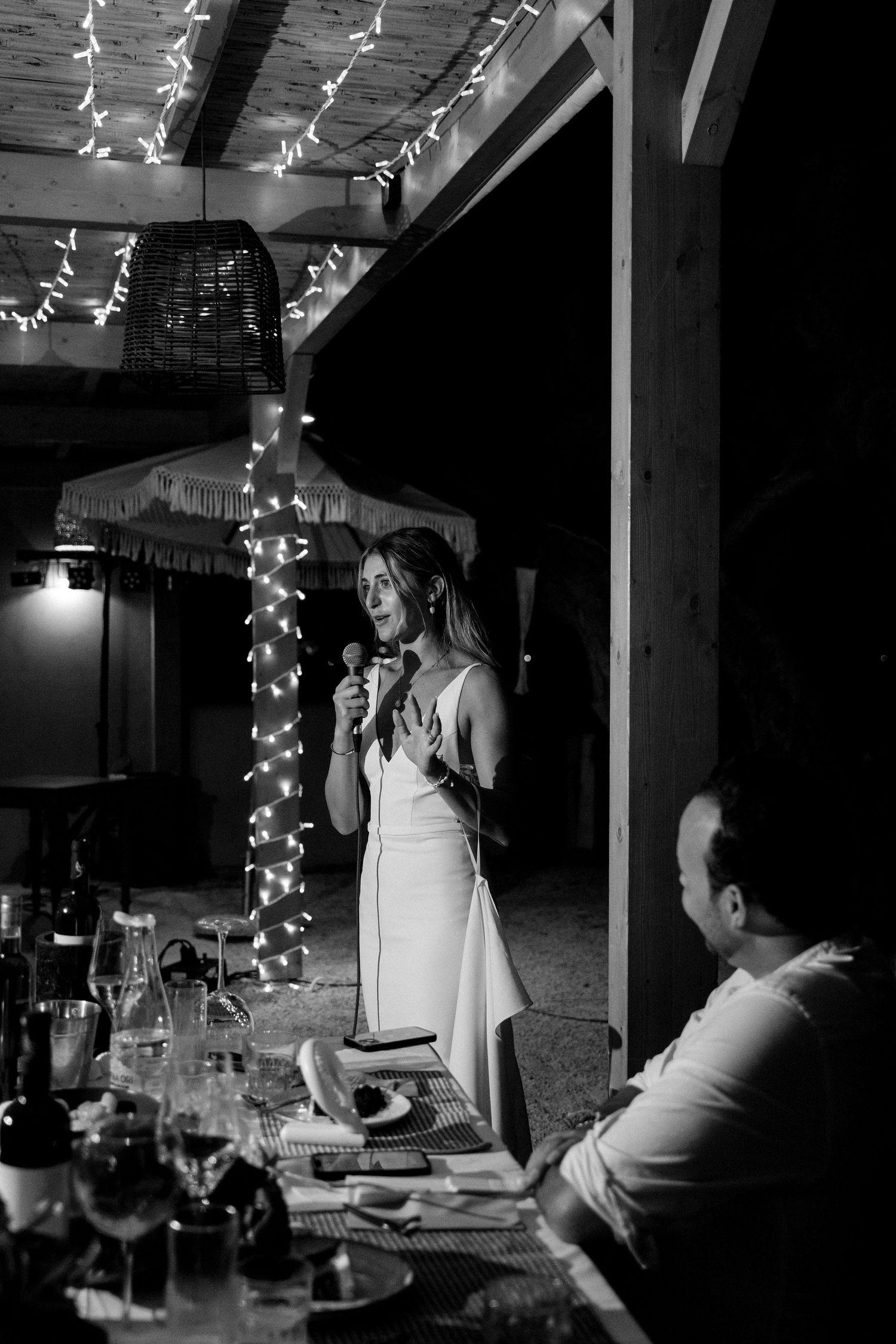 Black and white photo of wedding speech during an intimate reception with string lights and dinner table setting.